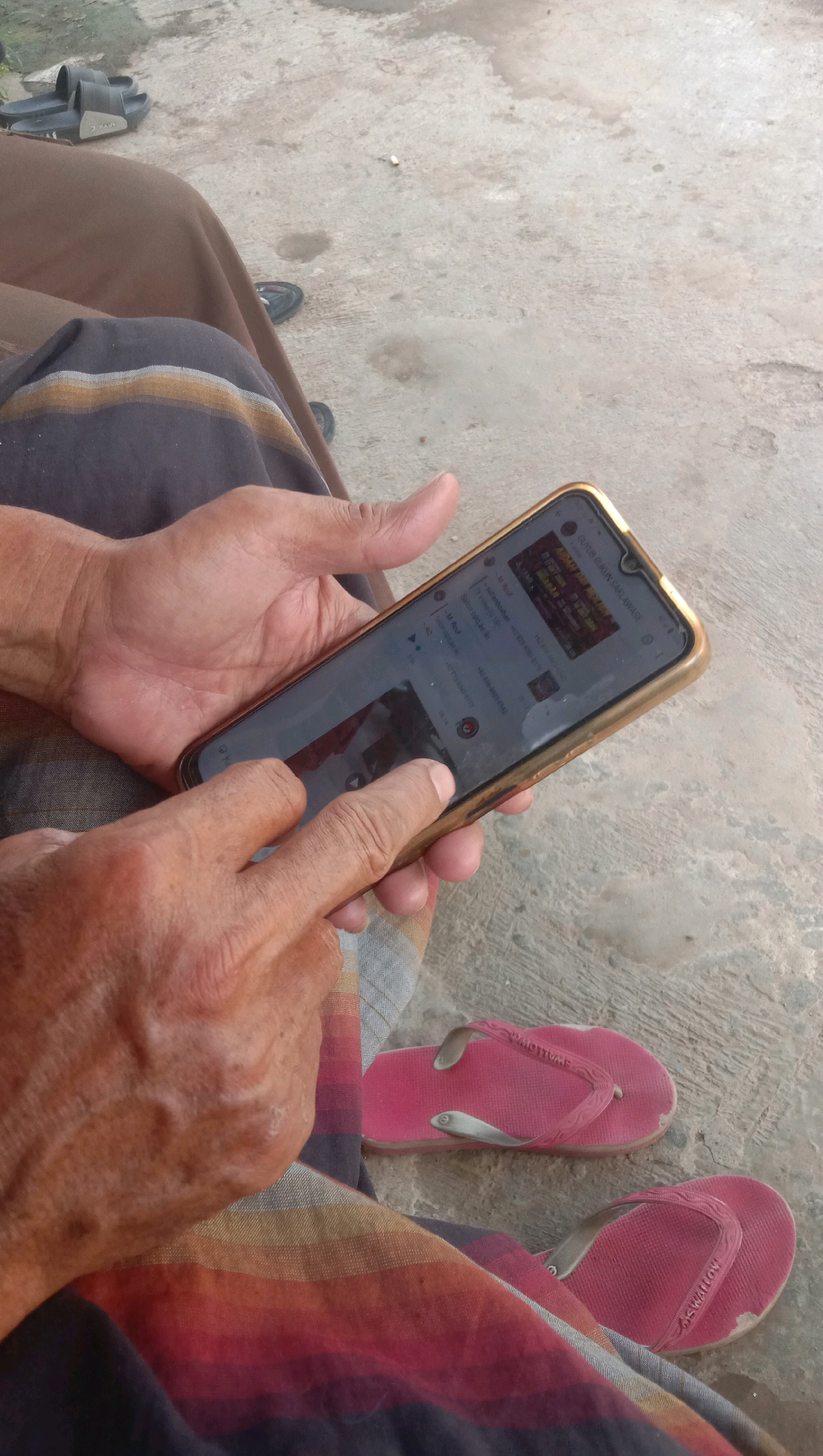 A close-up shot shows a person's hands holding a smartphone with a gold-colored case. The phone displays a chat application, with text messages and what appears to be a video thumbnail visible on the screen. The person's fingers are touching the screen, suggesting interaction with the device. The person is wearing a sarong with horizontal stripes in red, yellow, and black. Two pairs of pink flip-flops are visible on a concrete surface in the background. The scene appears to be outdoors, possibly in a residential area or public space in Karanganyar, Indonesia, indicated by the presence of local footwear and the general environmental elements.