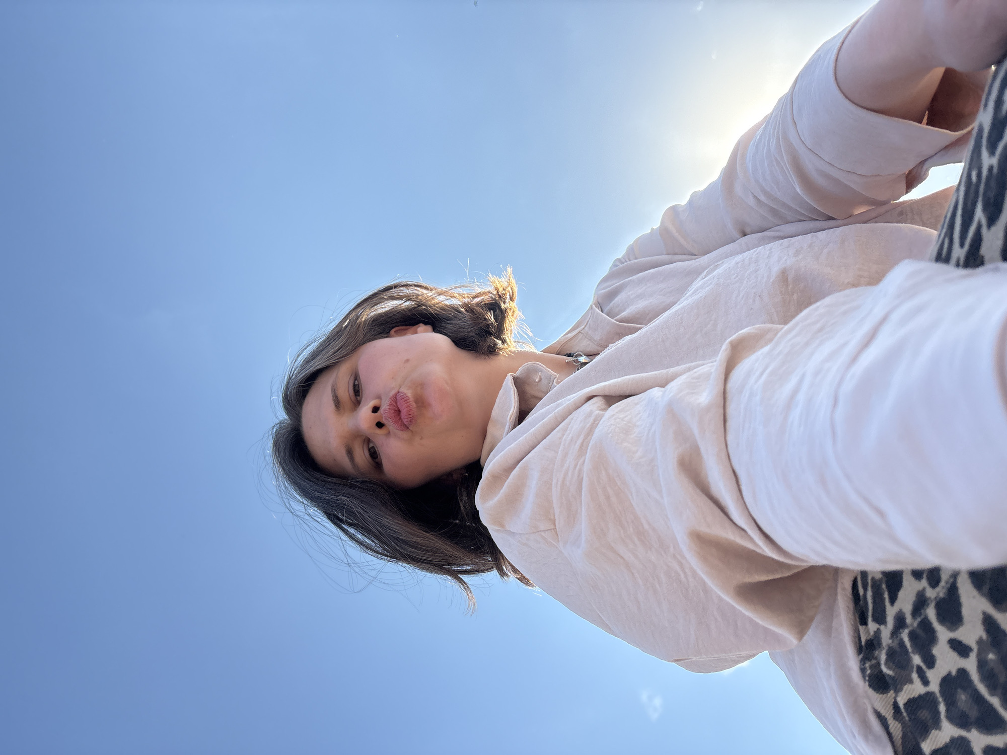 A woman with dark hair and a light complexion puckers her lips towards the camera in a selfie. She is wearing a light beige blouse and partially visible animal-print pants. The background is a clear blue sky with sunlight beaming from the upper right corner. It is difficult to ascertain the precise location in Chaikovsky, Russia, from this image alone.