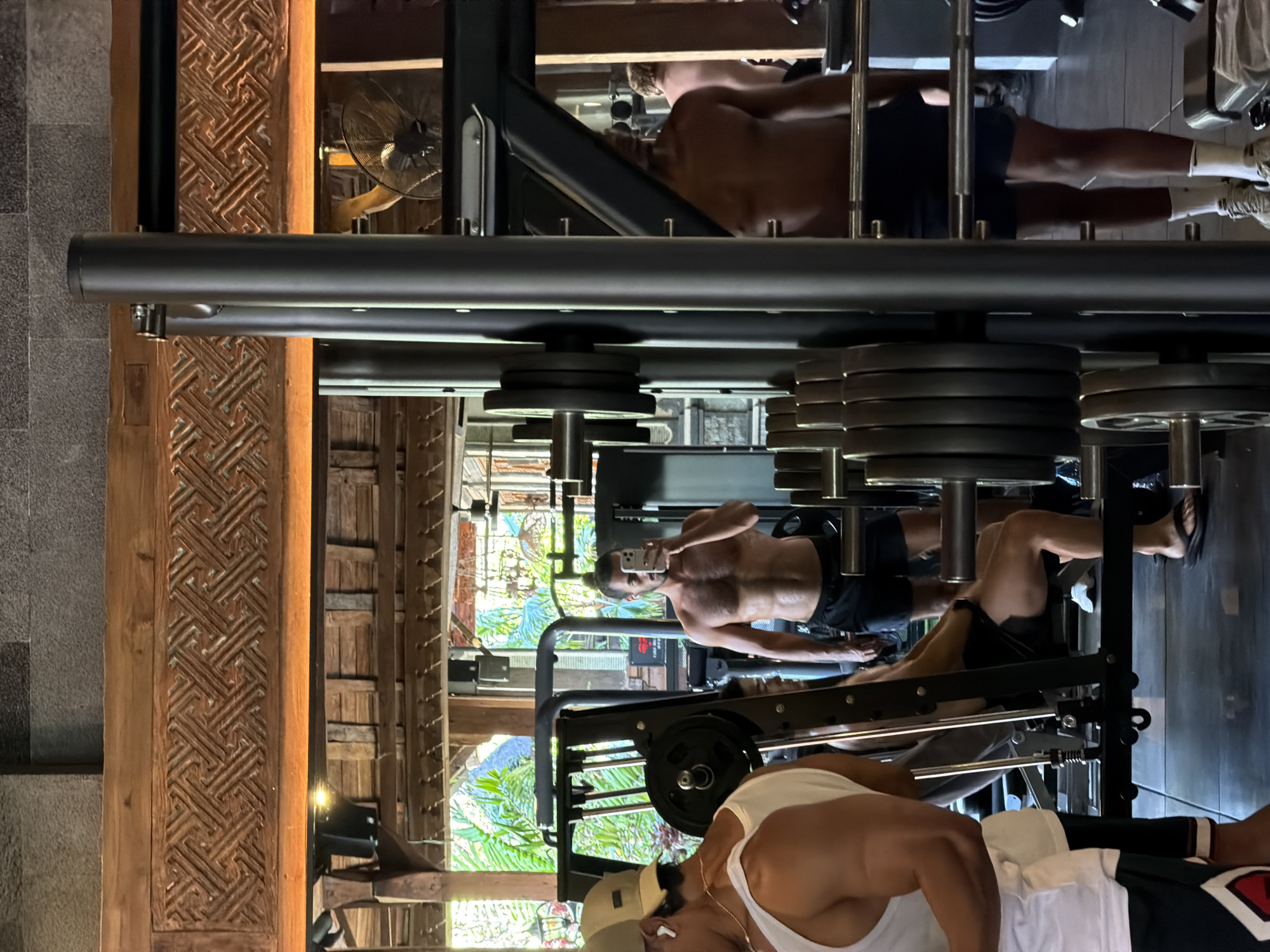 The image shows a gym interior, likely located in North Kuta, Indonesia, given the tropical foliage visible through a window or opening. The scene is dominated by gym equipment, including weight racks with stacked plates and a bench press machine.
The primary subjects are three men engaged in weightlifting activities. One man, shirtless and wearing black shorts, is performing a leg press on a machine. Another man, also shirtless and wearing black shorts, is positioned behind a weight rack, possibly preparing for a lift or resting. A third man, wearing a white tank top and a baseball cap, is partially visible in the foreground, with his arm and shoulder muscles prominent.
The overall aesthetic suggests a modern, well-equipped gym with some rustic elements, such as the intricately carved wooden pillars and textured stone walls visible in the background. The lighting appears to be a mix of artificial gym lighting and natural light, creating a slightly dramatic ambiance. The presence of lush greenery in the background suggests it is daytime, and possibly warm weather, consistent with the location in Indonesia. The focus on physical activity and the prominent display of muscles indicate a strong emphasis on fitness and bodybuilding within this setting. There is no visible text in the image.
