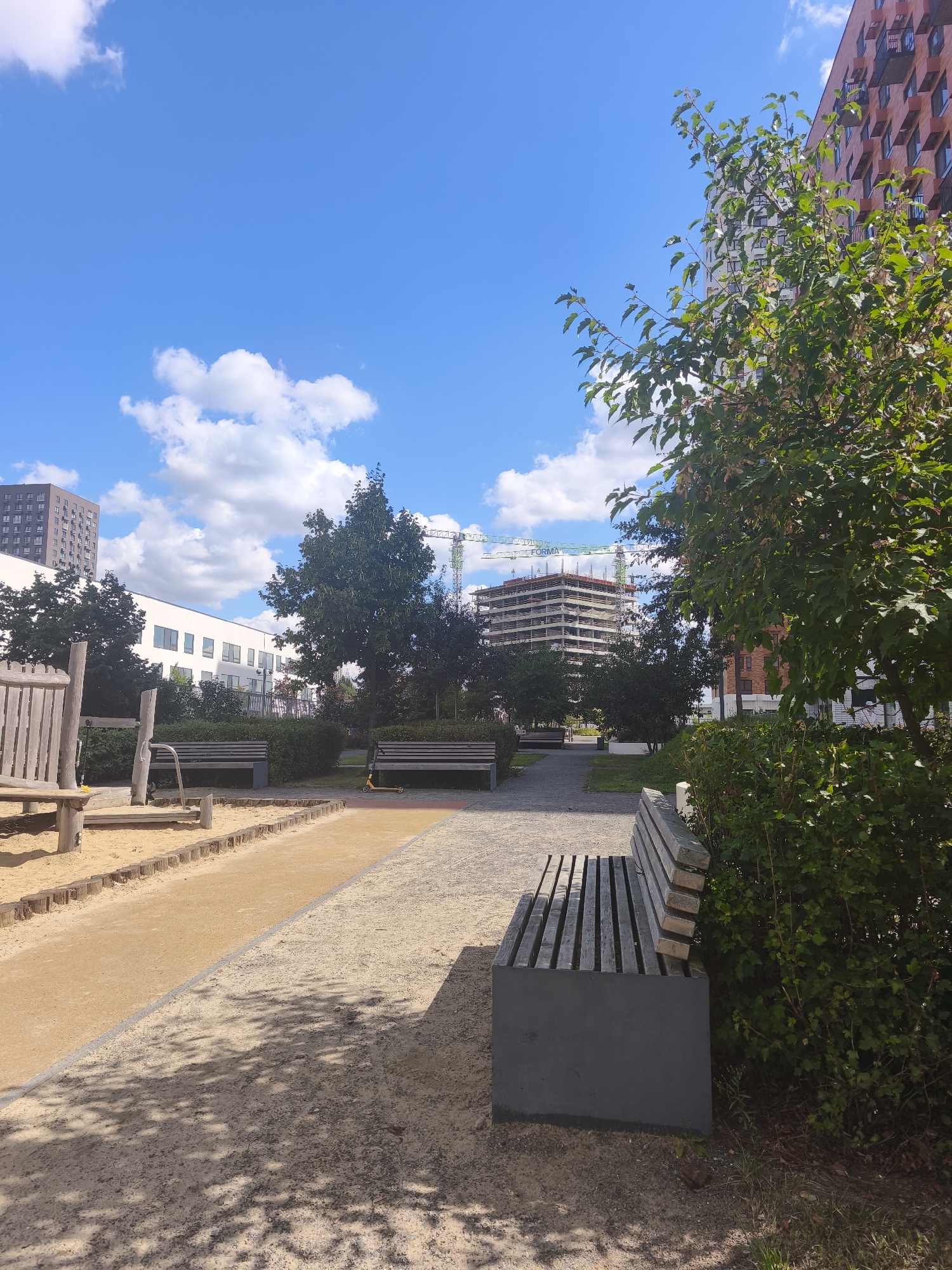This image depicts an outdoor scene in Moscow, Russia, likely on a bright, sunny day with a blue sky dotted with white clouds. In the foreground, a sand-filled playground area is visible on the left, with wooden play structures. A pathway made of gravel and colored paving stones runs through the middle ground, leading to several park benches. On the right side of the image, a modern, dark gray bench is situated next to a dense green bush.
In the background, several buildings are visible. A white building with multiple windows is on the left, and further back, a tall residential building with many windows can be seen. To the right, a large, partially constructed building stands, with a construction crane towering over it. Another modern building with a red brick facade and balconies is also present. The overall impression is one of a developing urban area with amenities for recreation.