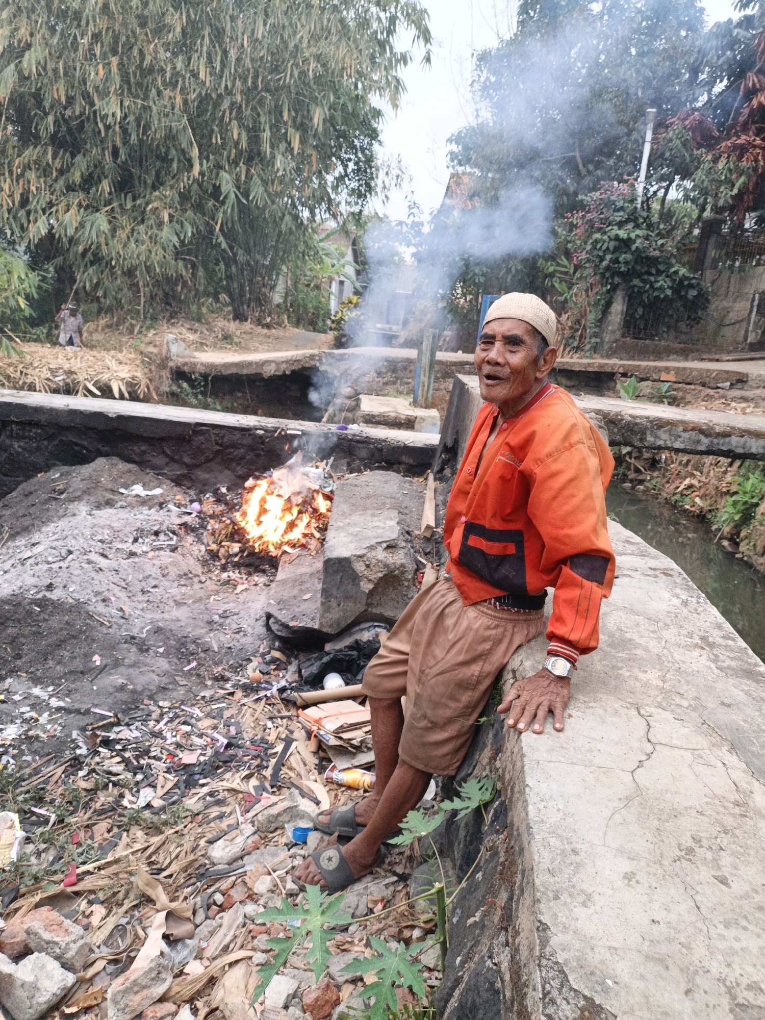 An elderly man with a light-colored skullcap and an orange jacket sits on a concrete structure next to a ditch. Smoke rises from a fire in the ditch, which contains burning trash. The man is looking towards the viewer and smiling. The surrounding environment is overgrown with vegetation, and there is evidence of litter and debris. A small stream or canal runs alongside the concrete structure. The scene appears to be located in a rural or semi-urban area, possibly in Mangli, Indonesia, given the context.