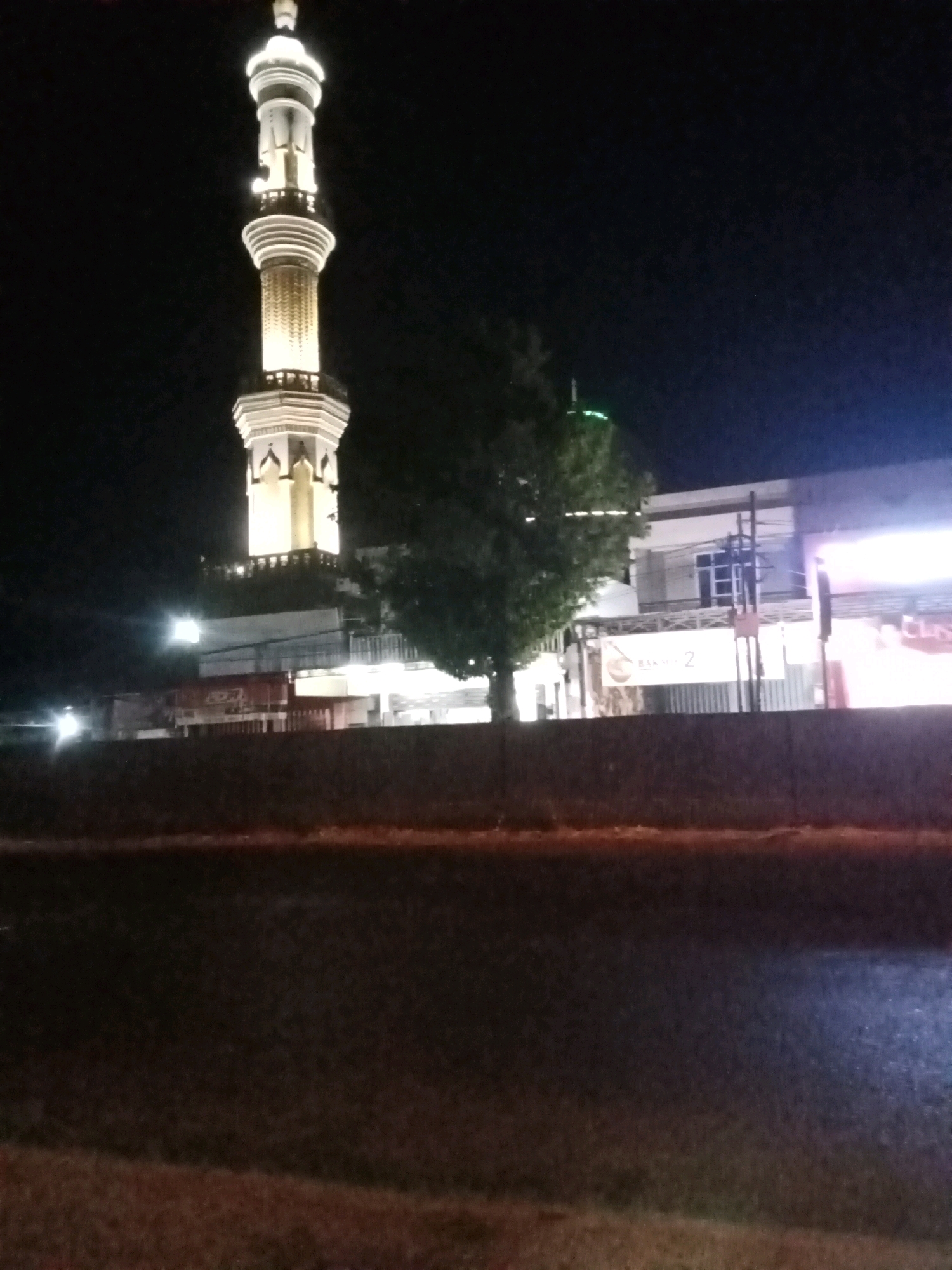 The image shows a minaret of a mosque illuminated at night, located in Peterongan, Indonesia. The minaret is tall and white with gold accents, and it is lit up, casting a warm glow against the dark sky. To the right of the minaret is a tree, and further to the right, a building with windows and several signs. One sign clearly visible in the center of the building reads "BAKSO 2". The foreground of the image is a dark road, possibly a street or highway, with some reflections from lights. The overall scene depicts a religious structure in an urban setting at night.