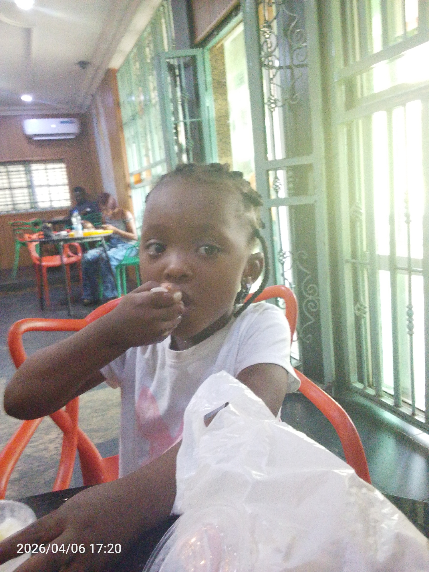 A young girl is sitting at a table in what appears to be a restaurant or food establishment in Lokoja, Nigeria. She is wearing a white t-shirt and eating something held in her hand. There is a white plastic bag on the table in front of her. In the background, other people are seated at tables, and there is a window with metal bars. An air conditioning unit is visible on the wall. The timestamp "2026/04/06 17:20" is visible in the bottom left corner.