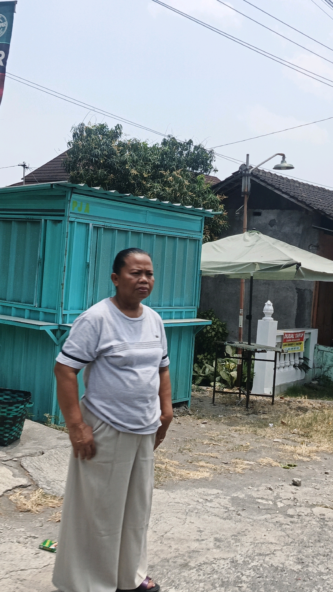 A medium shot captures an adult female standing outdoors in Selokaton, Indonesia. She is wearing a light gray t-shirt with a dark horizontal striped detail on the chest and loose-fitting, light beige pants. Her hair is pulled back. The background includes a teal-colored food stall, a residential building with a dark roof, an umbrella, a small table, a white decorative post, and a sign that reads "DIJUAL TANAH". Several power lines are visible overhead against a pale blue sky. The ground is composed of concrete and dirt with scattered dry vegetation. To the left of the woman, a dark woven basket is visible.