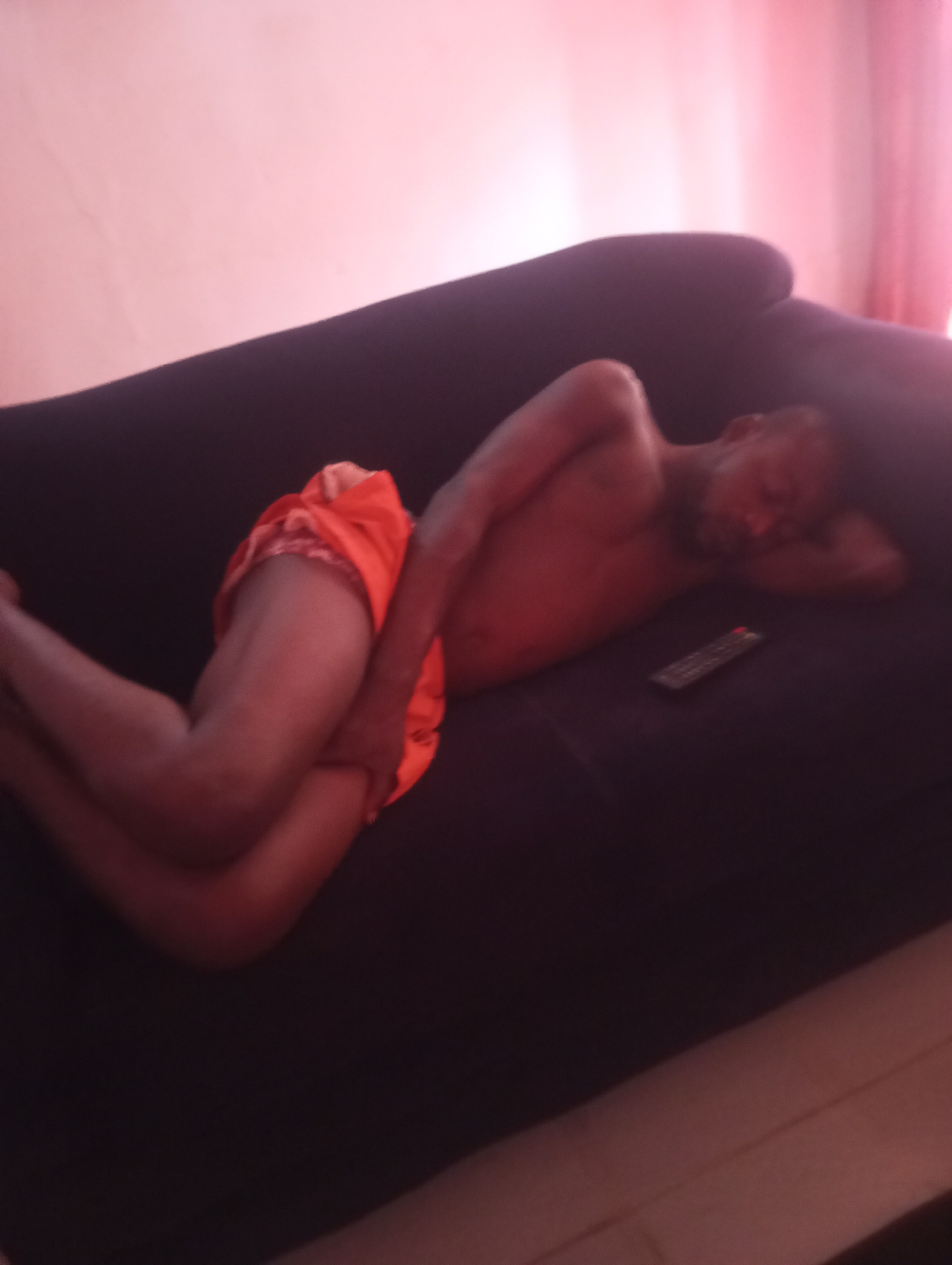 A man is lying on his side on a dark couch, asleep. He is shirtless and wearing orange shorts. His head is resting on his arm. A remote control is on the couch next to him. The background is a pink wall and a blurred pink curtain. The image appears to have been taken in a home in Yelwa, Nigeria.