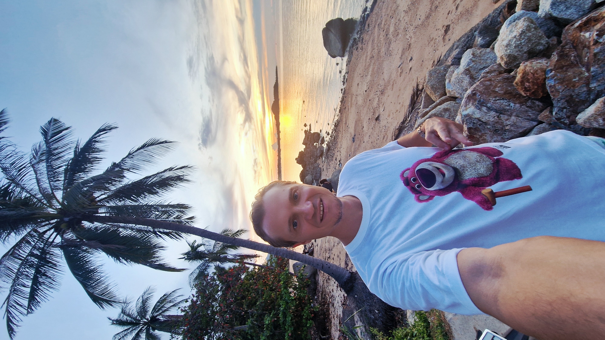A man stands on a beach at sunset, taking a selfie. He is wearing a white t-shirt with a pink cartoon bear character printed on it. He has short, light brown hair and is smiling. Behind him, a calm ocean reflects the warm hues of the setting sun. Palm trees line the sandy shore, with a few rocks scattered on the beach. In the distance, a faint silhouette of land is visible. The sky is a gradient of blues and oranges, with wispy clouds. The scene is located in Baan Bang Rak, Thailand, a coastal area known for its beautiful sunsets.