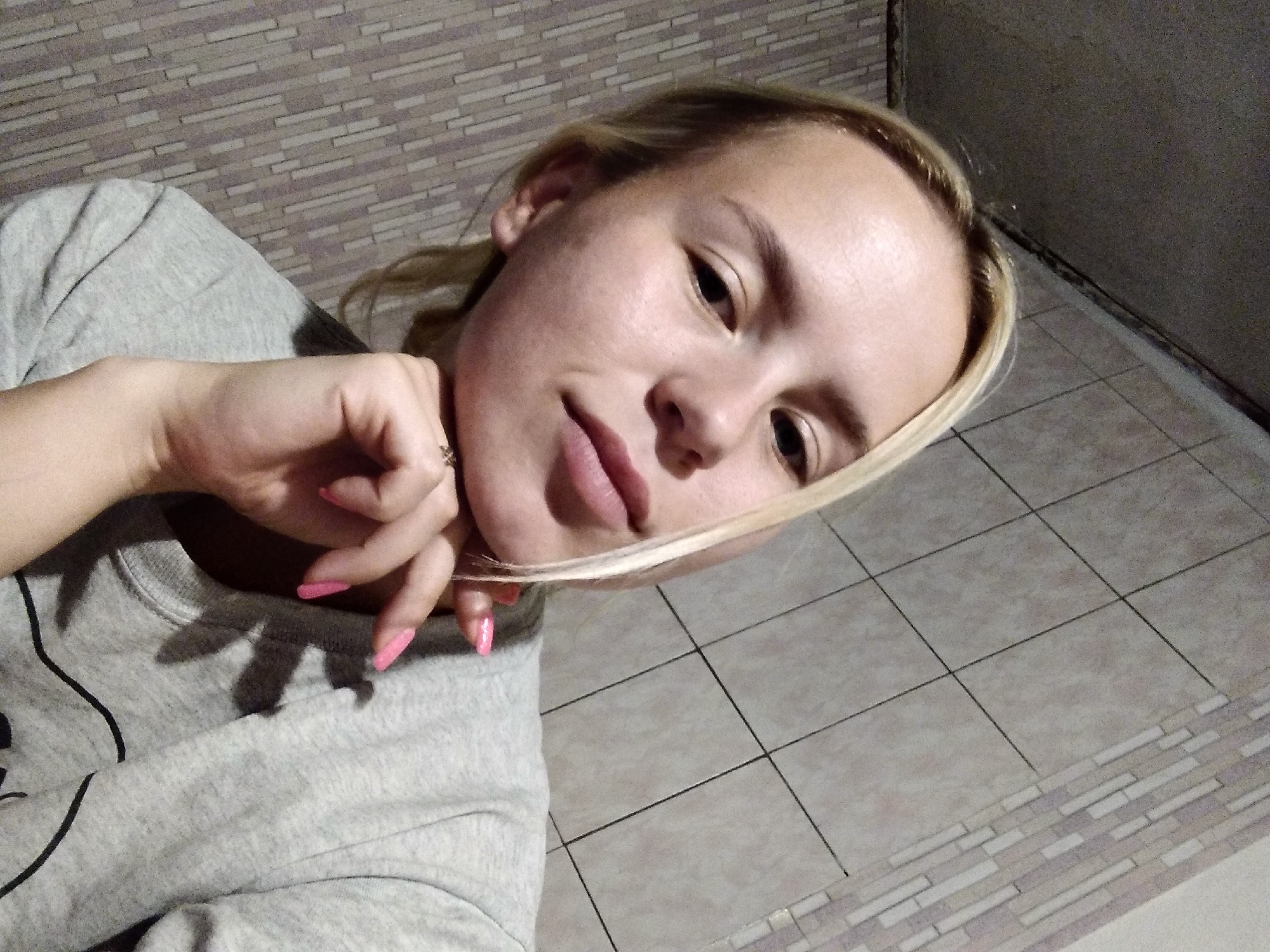 A young woman with blonde hair is captured in a close-up, slightly upward-angled selfie. She is wearing a grey t-shirt. Her left hand is resting on her chin, with her fingers framing her mouth. Her nails are painted a bright pink. The background appears to be a tiled wall with a pattern of rectangular tiles. The lighting is dim, casting shadows on her t-shirt.