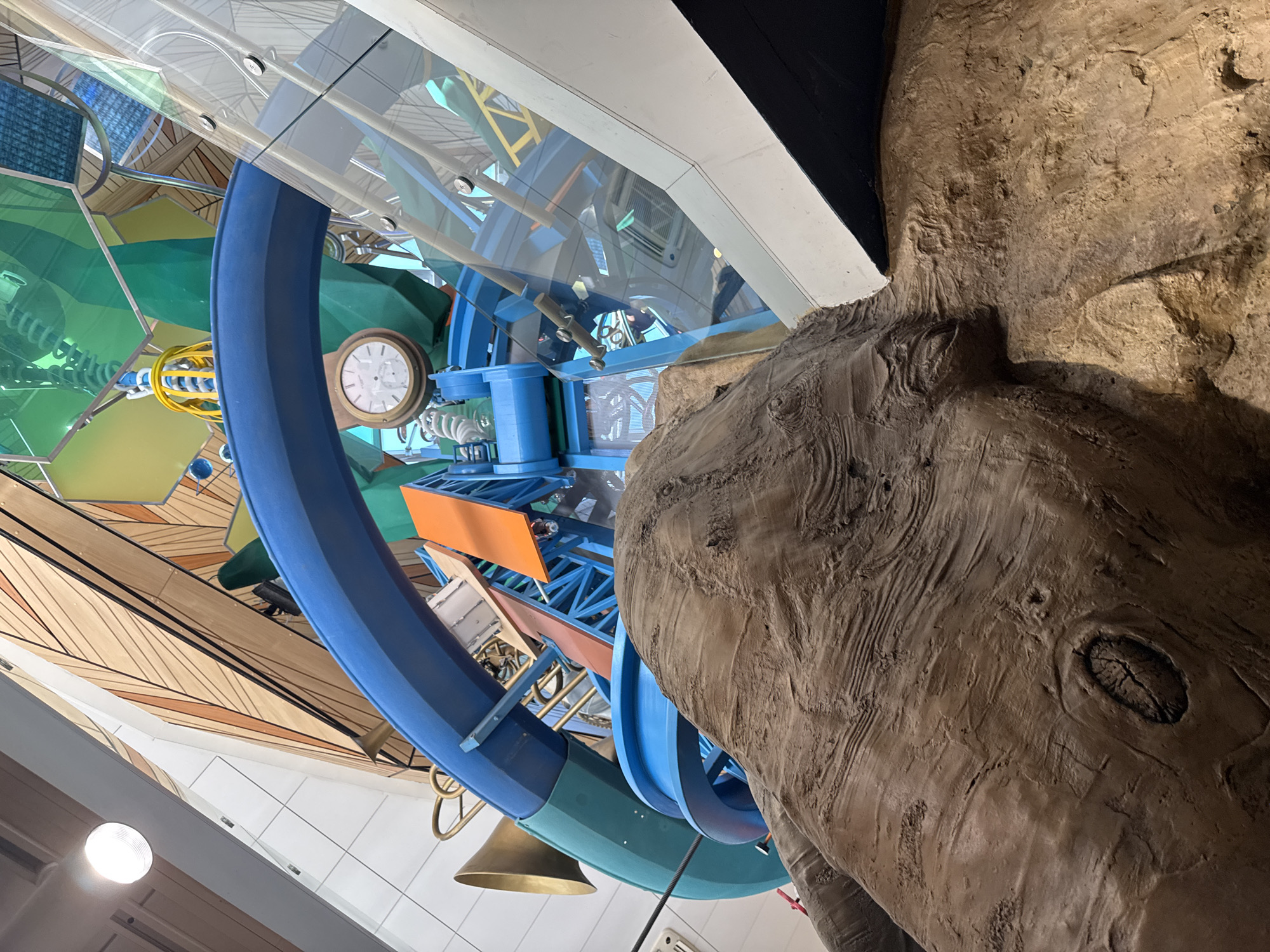The image depicts an interior scene featuring a complex, colorful installation. The installation includes elements such as blue circular tracks, various mechanical components, and sections with clocks and gears. The entire structure is partially enclosed by a clear, curved barrier, suggesting a protective display or enclosure. To the right of the installation, there's a large, organic-looking structure with a textured, brown surface, resembling either a tree trunk or a rock formation. The image is taken from a low angle, looking upwards, which causes some distortion of the perspective.
While specific landmarks or architectural details that definitively place the image in Sofia, Bulgaria are not apparent, the style of the installation and overall presentation is consistent with a museum or science center environment that may be found in such a city. Without further information, precise location identification remains speculative.