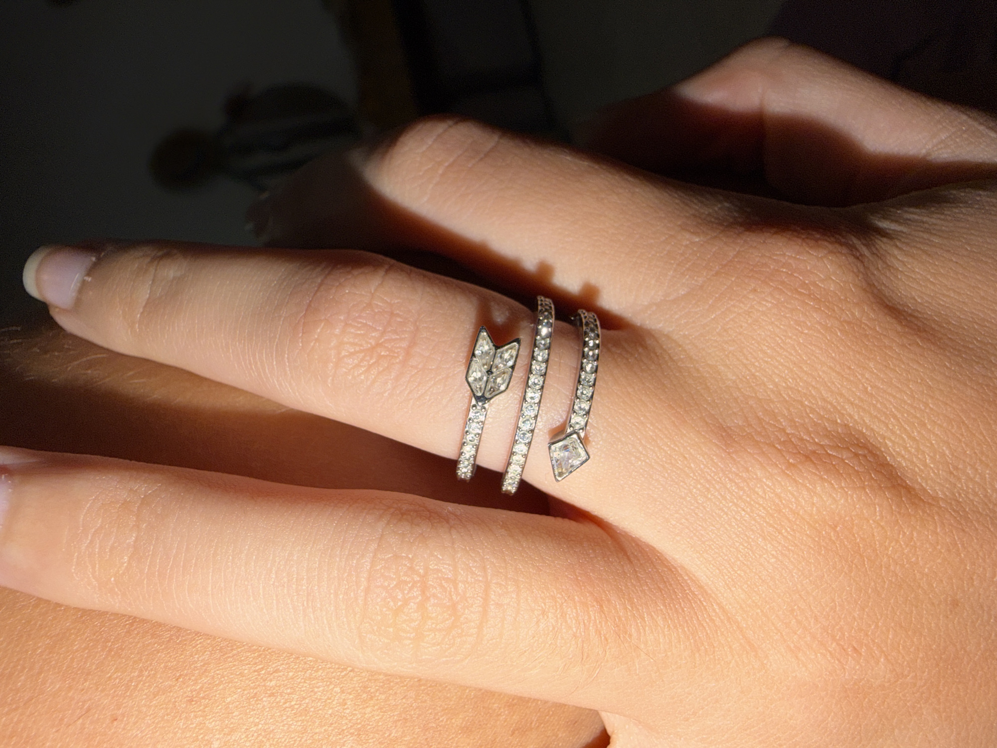 A close-up forensic photograph, captured in Bac Nha Trang Ward, Vietnam, focuses on a human hand adorned with a multi-band spiral ring. The hand exhibits a light-to-medium skin tone with visible skin texture, including fine lines and pores. The ring, worn on the ring finger, is silver-toned and features three distinct bands that wrap around the digit. Each band is encrusted with numerous small, round, brilliant-cut clear gemstones. The ring design incorporates an arrow motif: one end displays an arrowhead composed of three larger, square-cut clear gemstones, while the opposing end terminates in a single larger, square-cut clear gemstone, representing an arrow's fletching. The fingernail at the top of the finger is natural in appearance, short, and buffed. Direct, intense illumination originates from the upper-left, creating pronounced highlights on the ring's metallic surfaces and gemstones, and casting shadows that define the contours of the hand. The background is dark, heavily out-of-focus, and indistinct, with a blurred, vertical silhouette of a figure potentially wearing a striped garment visible on the far right.