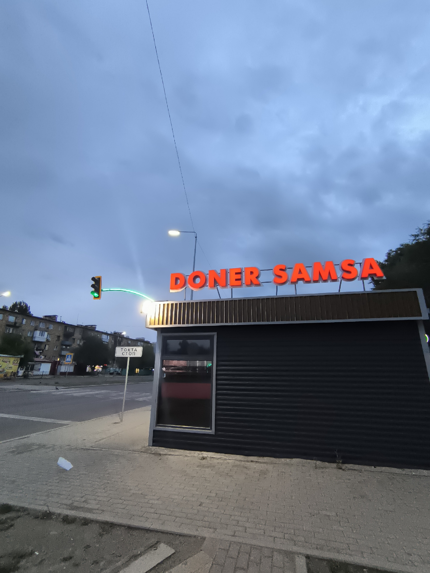 The image depicts an outdoor scene at dusk or early evening in Qarağandı, Kazakhstan. A modern, dark-colored food stall with illuminated orange lettering spelling "DONER SAMSA" dominates the foreground. The stall has a metallic, corrugated exterior and a wooden-paneled section above the storefront. To the left of the stall, a traffic light is visible, displaying a green light and a curved green signal extending towards the intersection. A white sign with black Cyrillic lettering, reading "TOCTA СТОП," meaning "Stop," is positioned near the traffic light. Across the street, apartment buildings and other smaller structures are visible. The sky is overcast with dark clouds. The ground in front of the stall is paved with gray interlocking bricks, and some debris, including a white piece of paper, is present on the pavement. The overall atmosphere suggests a commercial area in a city.