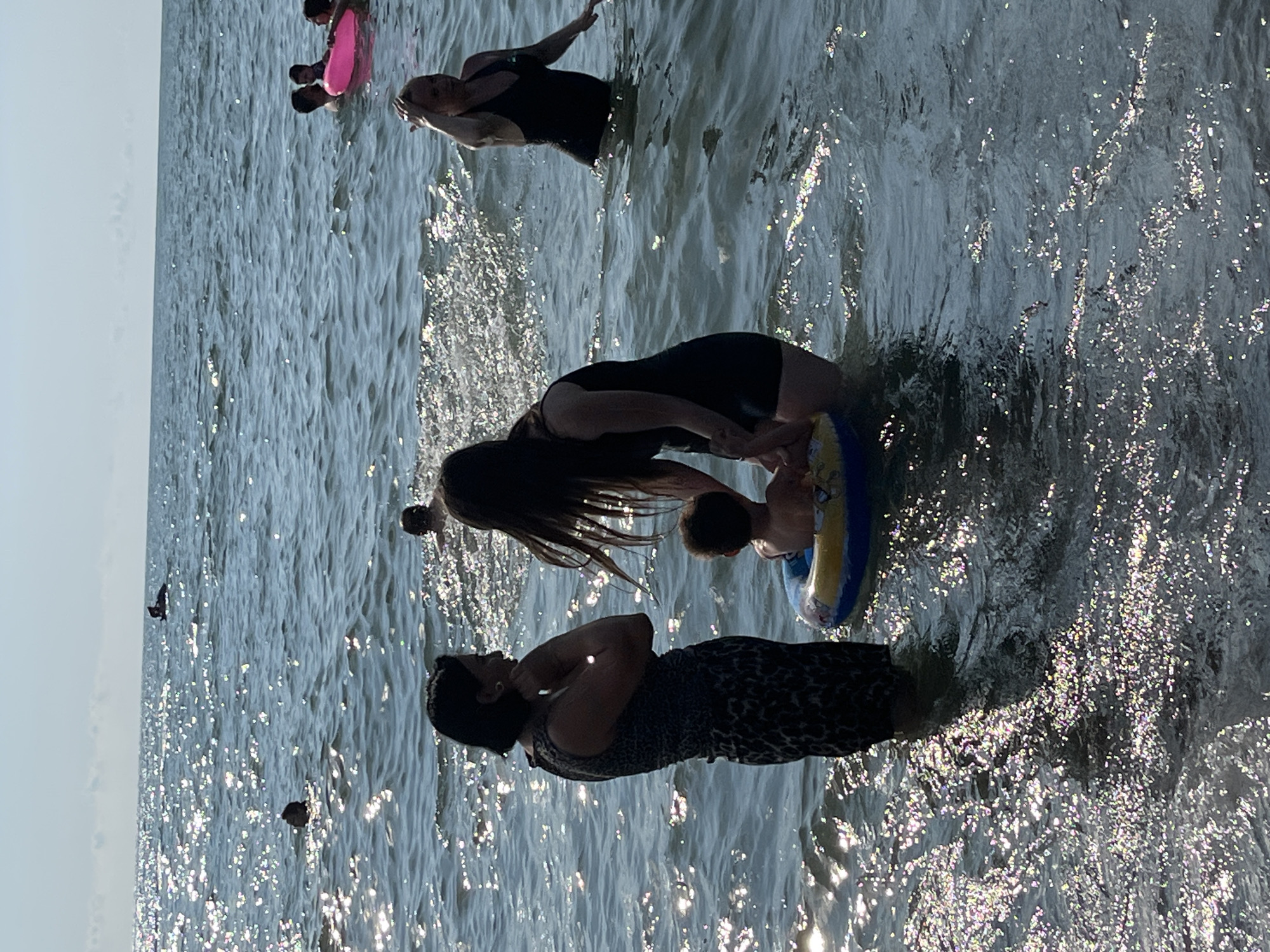 In Ureki, Georgia, on a sunny day, several people are enjoying the Black Sea. In the foreground, two women are in the water with a young child in a yellow and blue inflatable ring. One woman, on the left, wears a dark sleeveless top and a patterned skirt, facing away from the camera and looking towards the sea. The other woman, to her right, wears a black swimsuit and her hair is down. She is holding onto the child. Further out in the water, other individuals are visible swimming and interacting, including a person in a pink inflatable ring. The sea is choppy with sunlight glinting off the surface. The sky above is a light blue with some distant clouds.