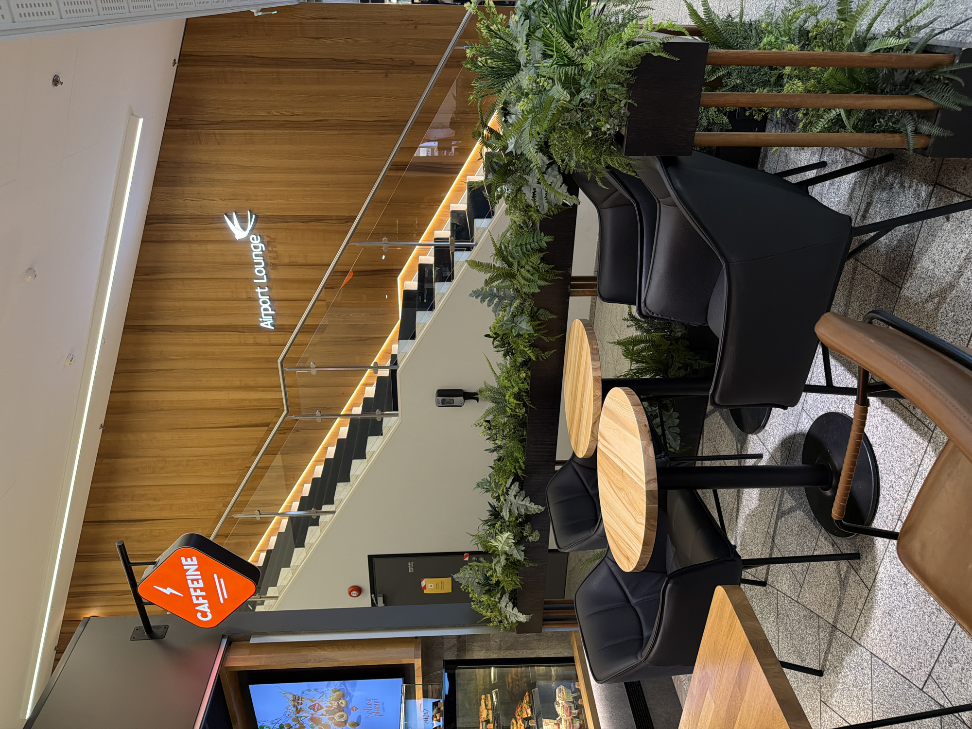An indoor airport lounge area in Tallinn, Estonia, features a modern seating arrangement with black leather chairs and light wood tables. A prominent orange diamond-shaped sign reading "CAFFEINE" with a lightning bolt symbol hangs on a dark gray wall to the left. Adjacent to the seating area, a staircase with black treads and risers is illuminated by a warm orange LED strip along its handrail. The walls are adorned with vertical wooden panels. A sign on the wall above the staircase reads "Airport Lounge" with a white bird-like logo. Lush green ferns and other plants are arranged in dark planters, softening the space. In the background, a display case showcases food items. The floor is a textured gray tile.