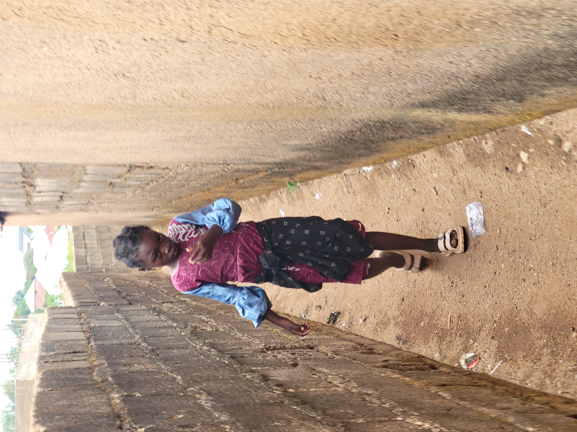 A young Black girl stands on a dirt path between a low brick wall and a road. She wears a red and white top with blue sleeves and a dark skirt tied at the waist. A pair of white sandals are on her feet. The surroundings appear to be an urban area, possibly on the outskirts of Jos, Nigeria, suggested by the style of the brick wall and the dusty environment. Discarded trash lies near her feet.