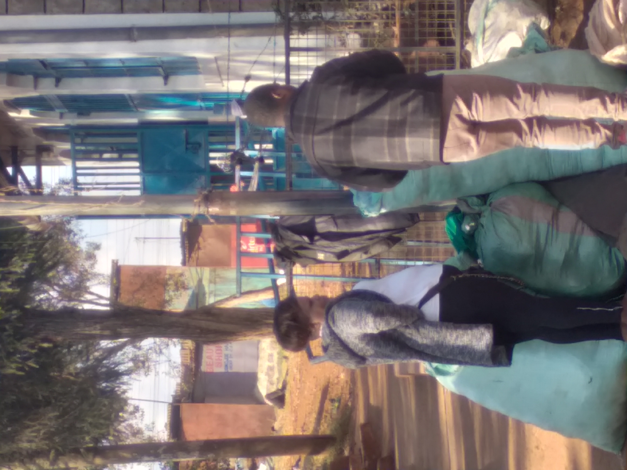 The image shows two individuals standing next to large sacks, possibly in Nyeri, Kenya. A woman in the foreground is wearing a grey hooded jacket over a white shirt and black pants, with her hands clasped in front of her. To her right, a man in a plaid jacket and light brown pants stands facing away from the camera. They are positioned on a dirt surface with large, light blue and green sacks to their left and right. In the background, a building with blue-trimmed windows and a blue metal gate is visible, along with trees and what appears to be a "BEAUTY SHOP" sign. The environment suggests an outdoor, possibly commercial or residential area with vegetation and structures typical of an urban setting in Kenya.
