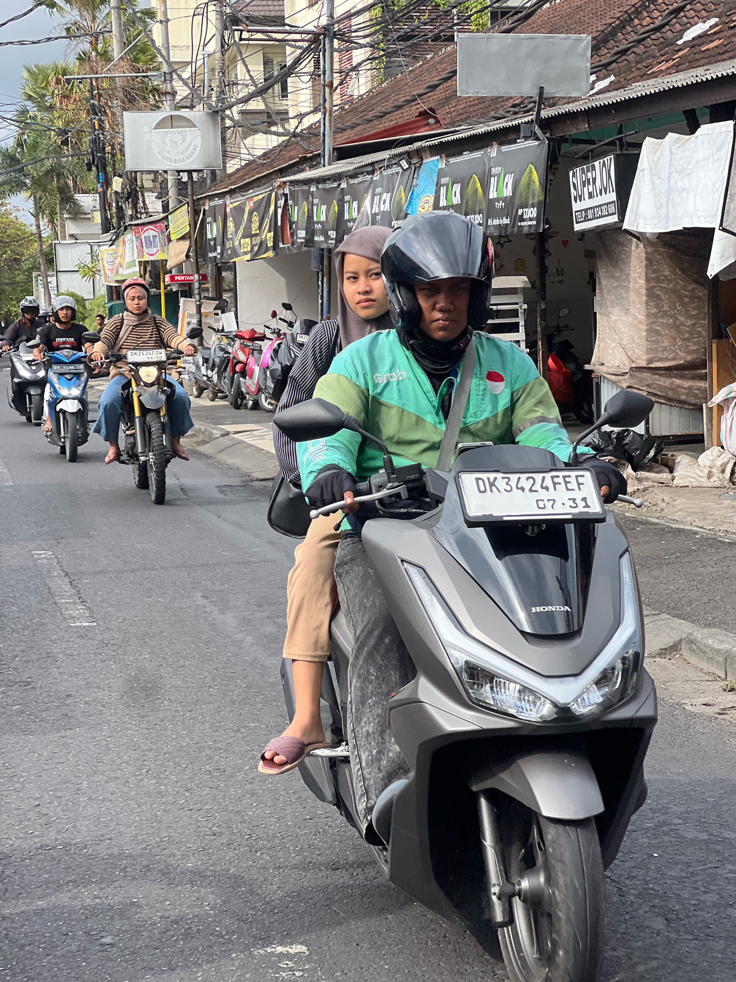 In Kuta, Indonesia, this image captures a street scene with multiple motorcycles in motion. The focal point is a gray Honda scooter with a license plate reading "DK3424FEF" and "07-31". The rider, wearing a black helmet and a turquoise and green jacket with the "Grab" logo, is in the foreground. A passenger behind the rider, wearing a gray hijab and a striped top, is also visible. To their left, another motorcycle carries a rider in a striped shirt and jeans. Further back, several other motorcycles and riders are present, suggesting moderate traffic on the asphalt road. The background features buildings with signs and numerous overhead electrical wires, typical of an urban Indonesian setting. Lush palm trees are visible behind the buildings, indicating a tropical climate. The overall scene depicts a busy street in Kuta.