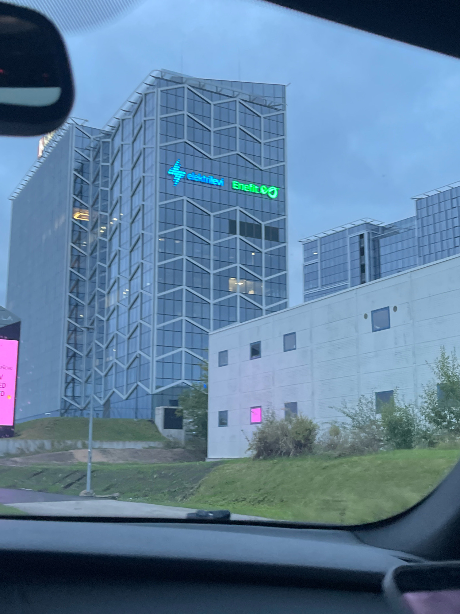 In Tallinn, Estonia, the image captures a segment of the Enerlift building, distinguished by its glass facade and a prominent green logo featuring a stylized cross and the text "Enerlift". The building is situated in an urban environment, with a contemporary architectural style. In the foreground, a section of a car's interior is visible, including the dashboard and part of the rearview mirror, indicating the photograph was taken from inside a vehicle. Adjacent to the Enerlift building, a lower, lighter-colored structure with a facade of white panels and multiple rectangular windows is also present. One of these windows emits a pinkish glow. The sky above is overcast, suggesting a cloudy day or evening. Green grass and some trees surround the base of the buildings, contributing to the urban landscaping.