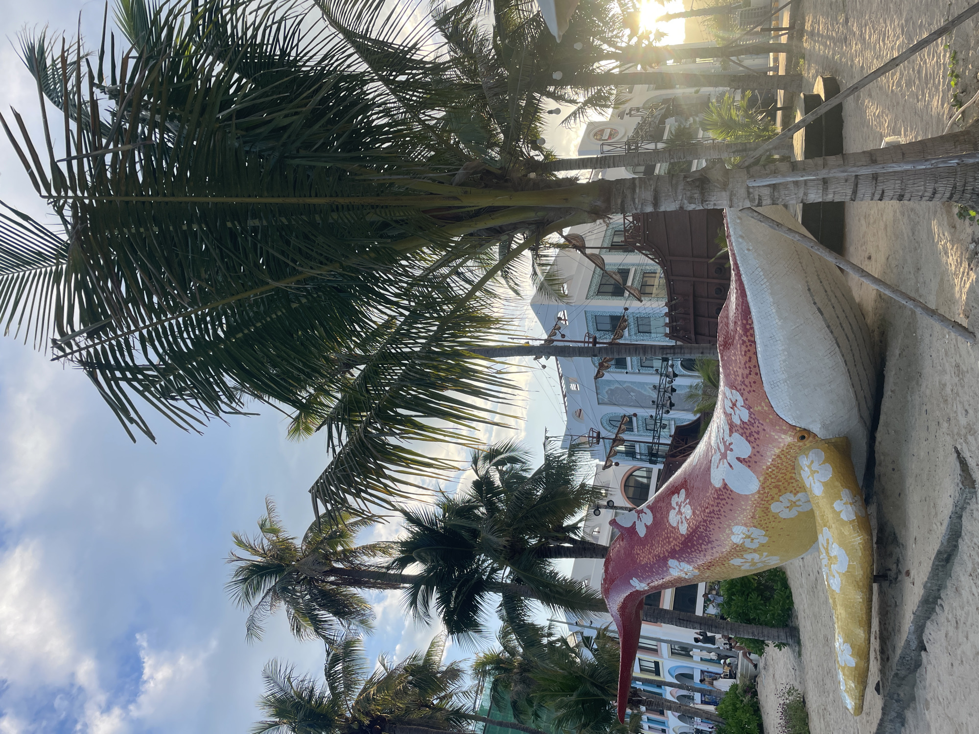 The image shows a tropical beach scene in Bac Nha Trang Ward, Vietnam. Palm trees dominate the left side of the frame, reaching up towards a partly cloudy sky. The sun is shining brightly, partially obscured by the leaves of the trees. A colorful, mosaic-covered animal sculpture, possibly a pelican, sits on the sandy ground, its head and beak pointing left. The background features buildings, possibly hotels or resorts, with multiple stories and windows. Wooden structures with thatched roofs are also visible amidst the buildings and trees. The overall scene suggests a resort area with a beach and tropical vegetation.