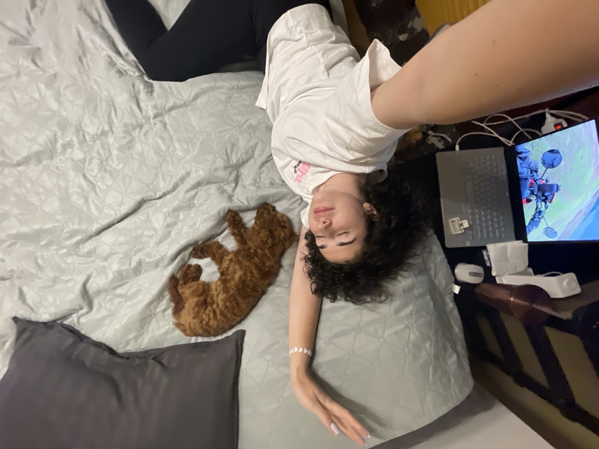 A woman is lying on a bed with gray sheets and a gray pillow. She is wearing black pants and a white t-shirt. A brown poodle is lying next to her. A laptop is open on a table next to the bed, with a motorcycle image displayed on the screen. A computer mouse and other objects are on the table. The location is not discernible from the image.
