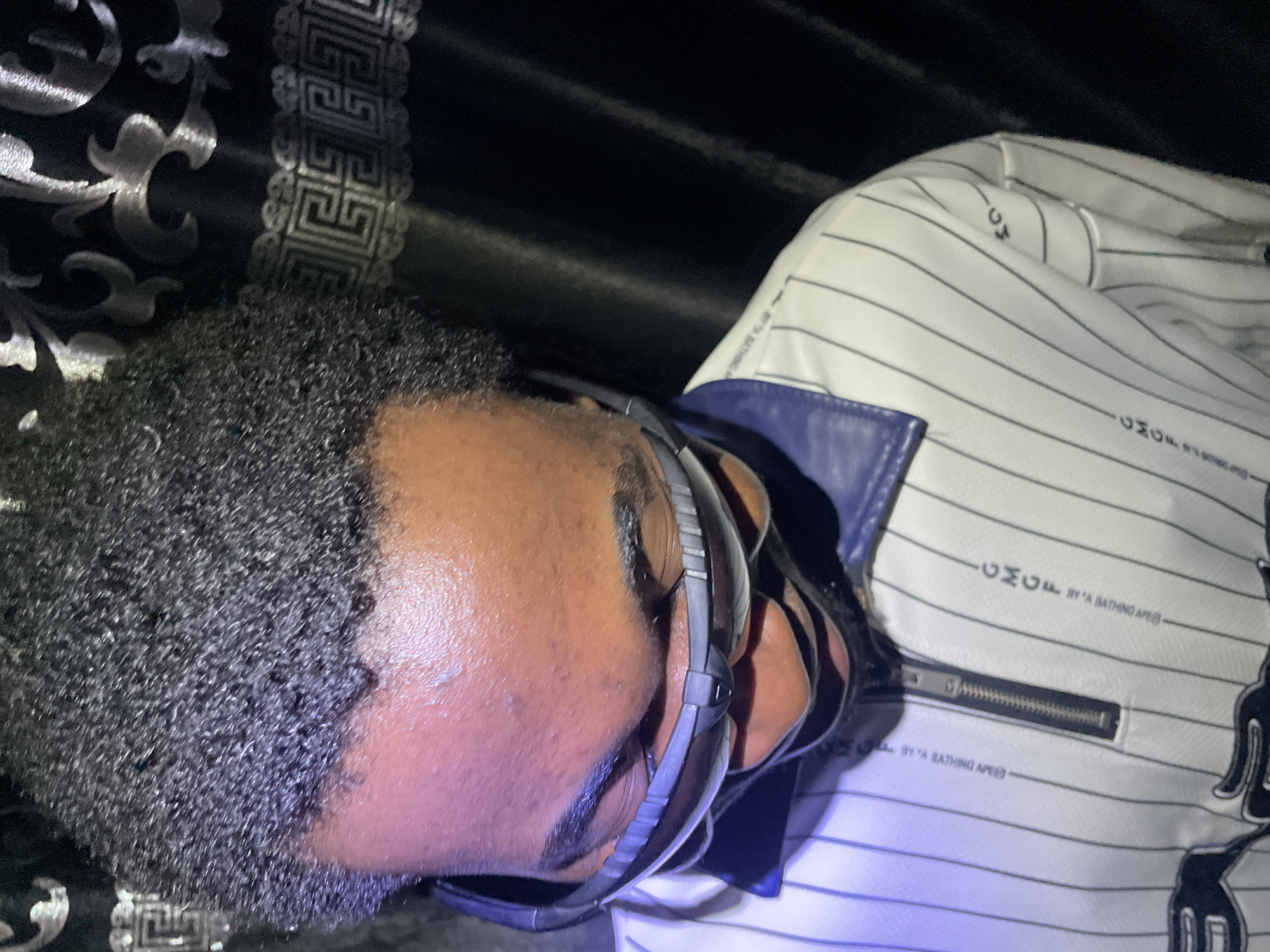 A man with short, curly black hair and dark skin is wearing gray framed glasses. He is wearing a white and blue jacket that has black vertical stripes and the words "CMCF BY A BATHING APE" printed on it. The jacket has a silver zipper visible. The background is a dark fabric with a silver and black pattern. The location in Lokoja, Nigeria is not discernible from this image.
