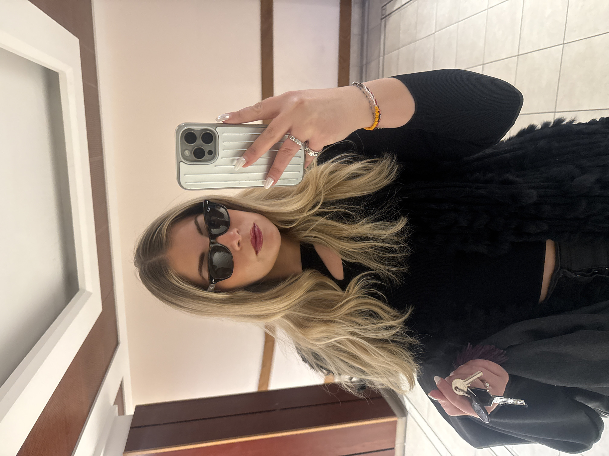 A woman takes a selfie in what appears to be a bathroom. She is wearing sunglasses, dark clothing, a bracelet, and rings. She is holding a cell phone in one hand and keys in the other. The background includes a door frame and tiled flooring. There's no information in the image to indicate the location is Paris, France.