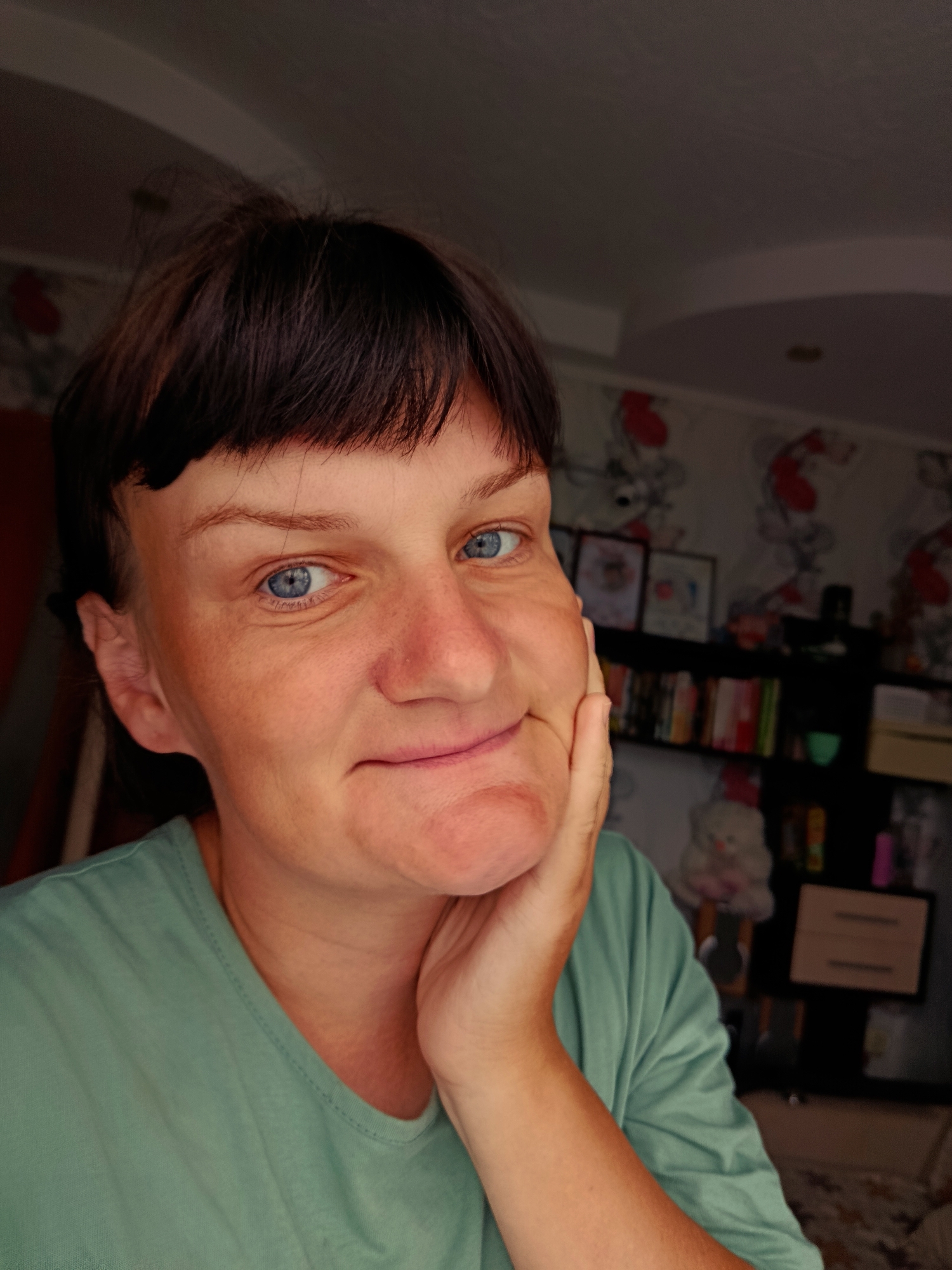 The image shows a close-up, selfie-style shot of a fair-skinned woman with dark brown hair and blue eyes. She is wearing a light green t-shirt. Her right hand is placed on her right cheek, and she is looking directly at the camera with a slight smile. The background appears to be an indoor setting, possibly a room in an apartment, with patterned wallpaper and a shelf filled with books and other items, including a stuffed animal. There are no visible indications of the location being in Фоки, Russia.