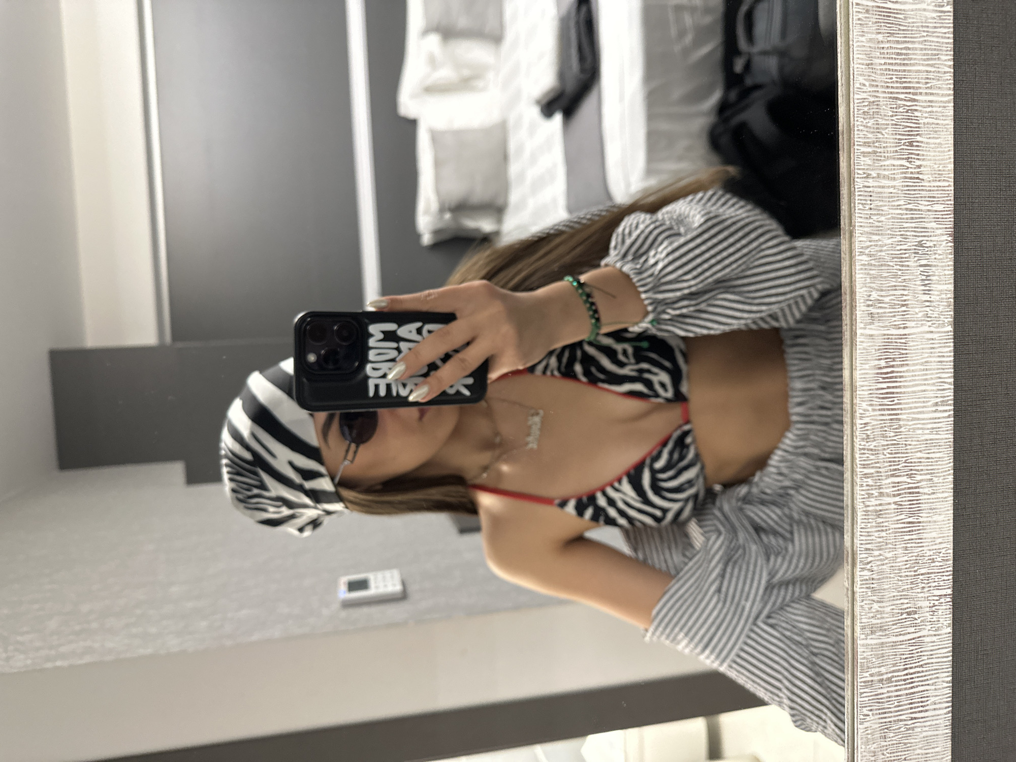 A young woman is reflected in a mirror, taking a selfie. She is wearing a zebra-print bikini top, a striped unbuttoned shirt, sunglasses, and a zebra-print bandana. She is also wearing a necklace and a bracelet. The background appears to be a hotel room, with a bed visible behind her. The location is Ofrynio Beach, Greece.