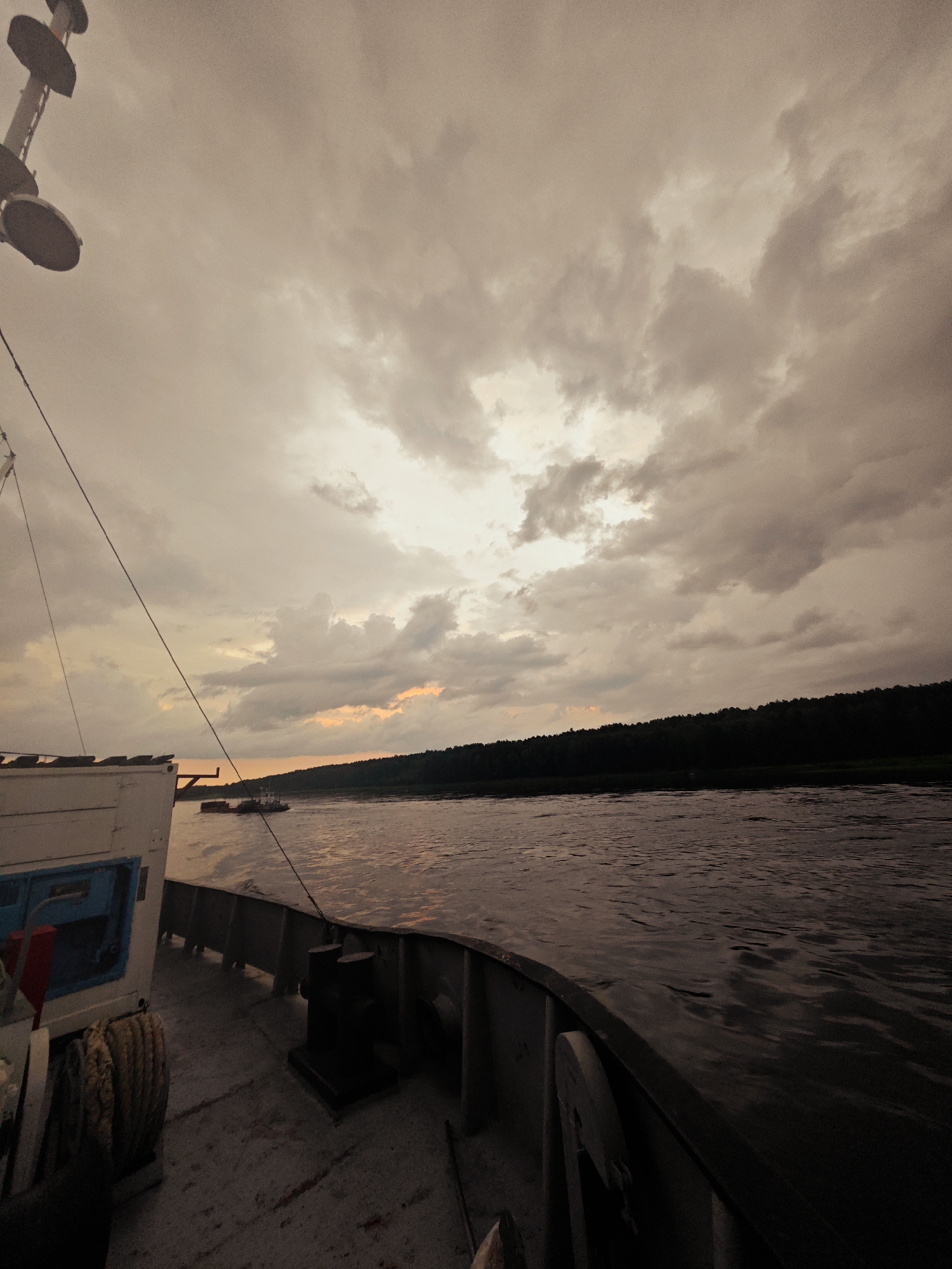 This media file depicts a view from the deck of a boat sailing on a river, likely in Kazachinskoe, Russia. The time of day appears to be either dawn or dusk, as suggested by the warm, muted tones of the sky and the soft lighting. The sky is overcast with dramatic clouds, creating a somber or contemplative mood.
The foreground of the image shows a portion of the boat's deck, with a coil of thick rope prominently displayed to the left and a dark bollard situated in the center. A white structure with a blue opening is also visible on the deck. A dark rope stretches diagonally across the frame, connecting to unseen parts of the boat.
In the mid-ground, the river surface is textured with ripples, reflecting the muted light. Further in the distance, another boat, possibly a tugboat, is visible. The far bank of the river is lined with a dense, dark forest, creating a stark silhouette against the sky.
The overall impression is one of solitude and the quiet movement of a vessel through a natural landscape under a dramatic sky. There is no visible text in the image.