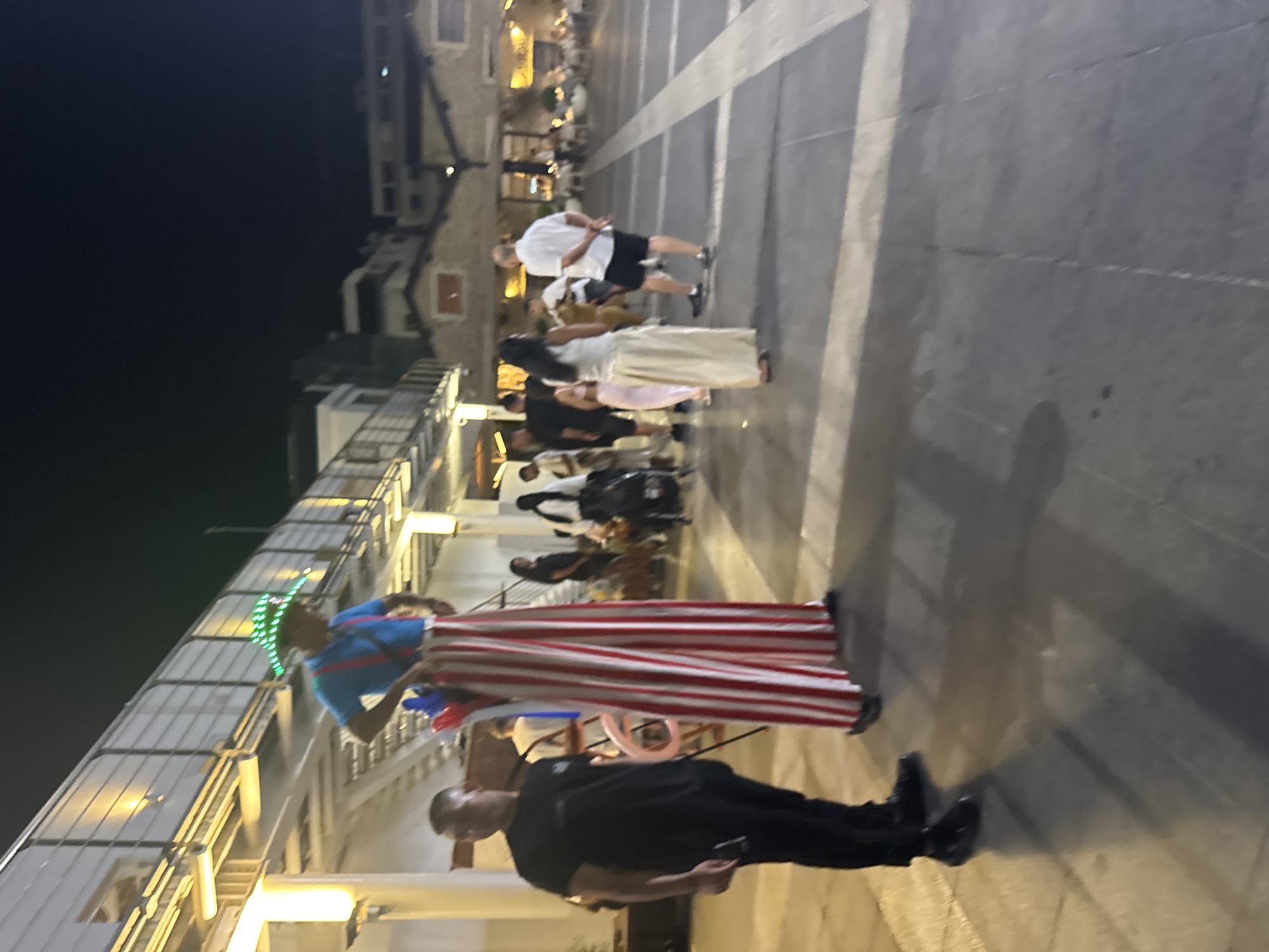 At night, a male stilt walker wearing a blue shirt, red suspenders, and red and white striped pants with a green LED hat is standing in a plaza in Limassol, Cyprus. He is holding some red and blue balloons. A man in a black t-shirt and black pants is standing to his left, looking towards the stilt walker. Several other people are visible in the background, some walking and others seated at tables in what appears to be an outdoor dining area. The plaza has a tiled floor and a raised walkway with railings and lights runs along the left side of the image, suggesting a promenade or entertainment area near the coast. Buildings are visible in the background, lit up at night.