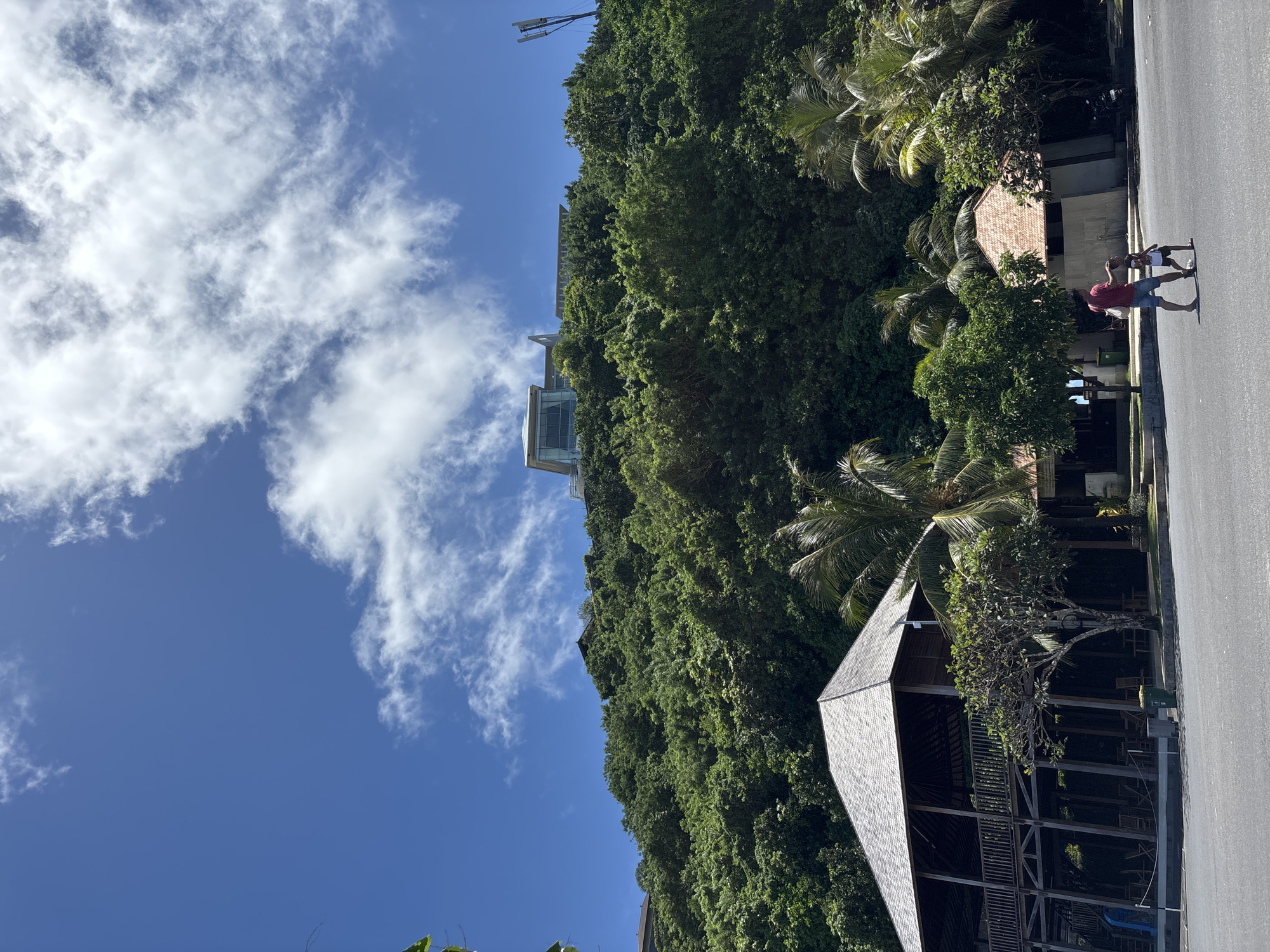 The image shows a view in Ungasan, Indonesia. The sky is blue with scattered white clouds. Lush green vegetation covers the landscape, including trees and dense foliage. A modern, possibly multi-story building is partially visible amidst the trees. In the foreground is a wooden structure with a peaked roof. A man and child are seen on the street walking near other houses. The street appears to be paved.