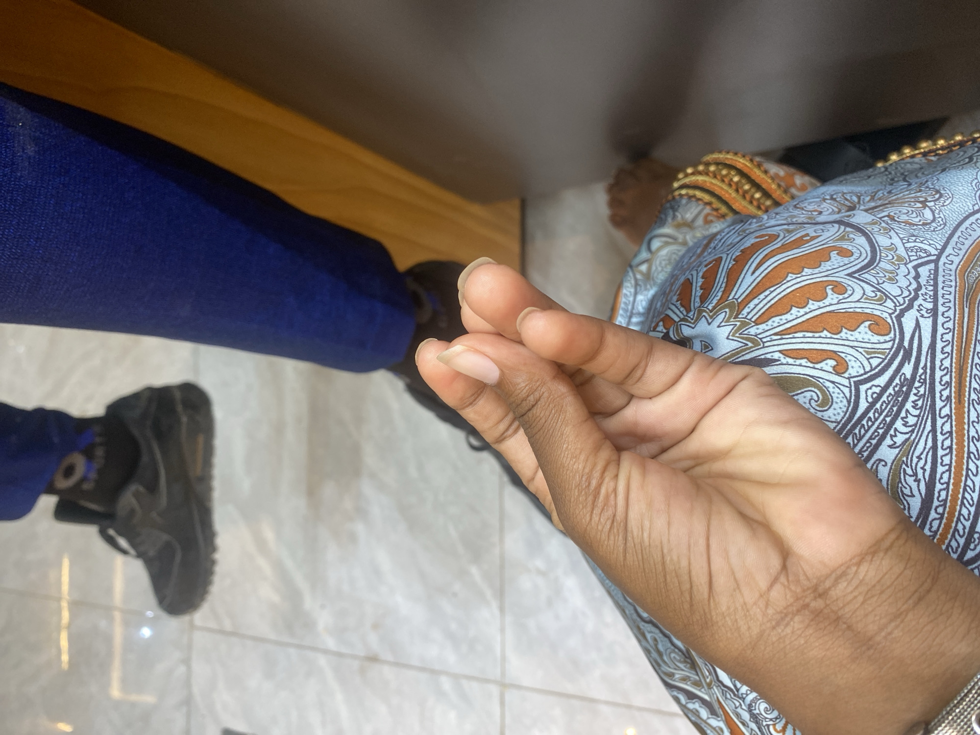 The image shows a scene indoors, likely on a tiled floor. Two individuals are present, partially visible. The individual on the left wears dark grey sneakers, blue pants and is standing near what appears to be the edge of a wooden furniture piece. The individual on the right wears a patterned blue and brown garment with decorative gold balls along the hem. Their hand is prominently displayed in the foreground. The location cannot be determined from the image.