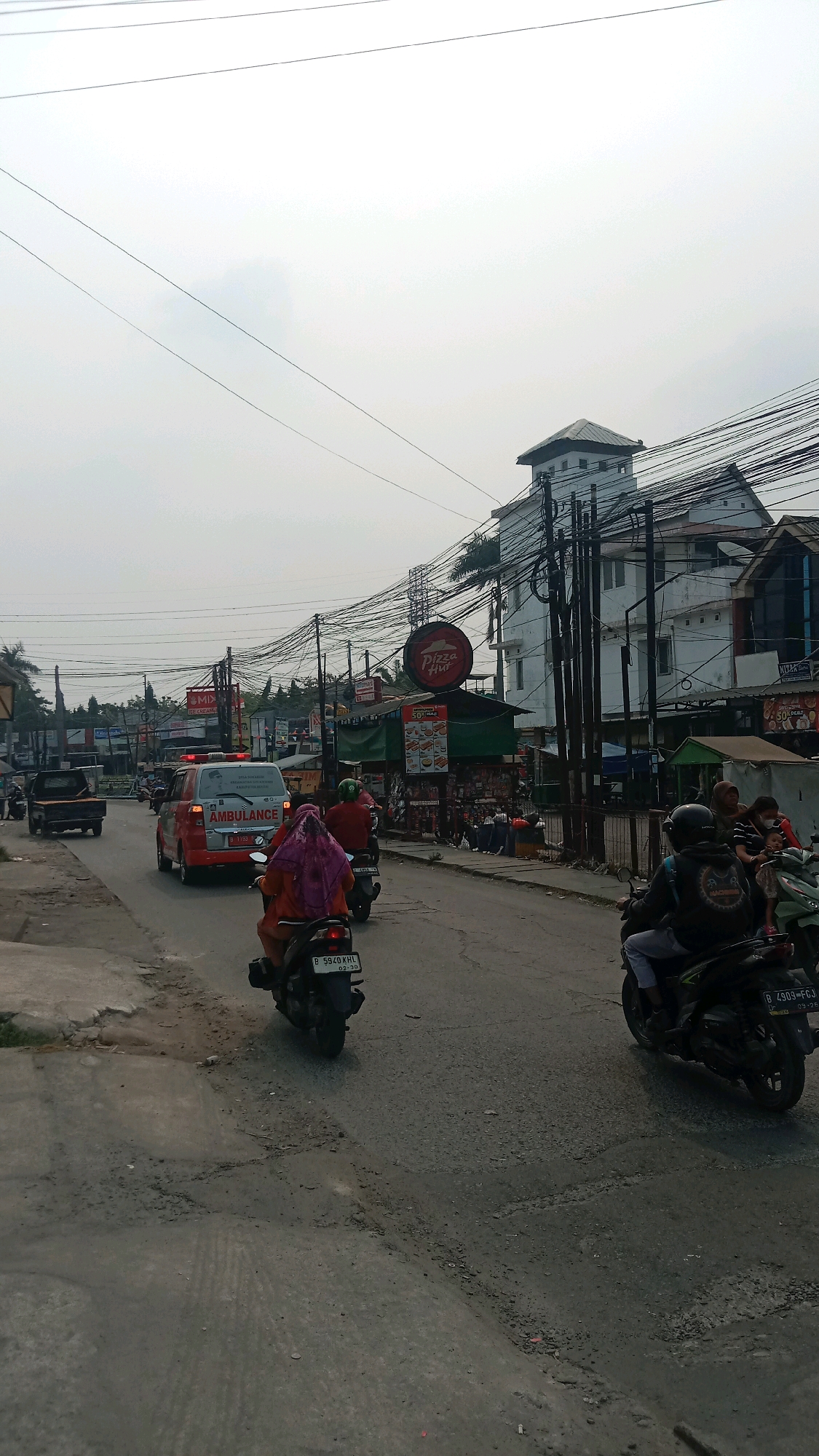The image depicts a street scene in Wanasari, Indonesia, under an overcast sky. The road is paved with asphalt and shows signs of wear and tear, with visible potholes and cracks. On the right side of the road, a two-story building with white walls and a dark-tiled roof is prominent, with numerous electrical wires crisscrossing above it. A red circular Pizza Hut sign is affixed to the building's facade. Shops with green awnings are situated below the Pizza Hut, displaying menus or advertisements. Several people are present, some on motorcycles and others on foot, interacting within the commercial area.
On the road, traffic is moving. An ambulance, white with red detailing and the word "AMBULANCE" clearly visible, is a central focus, traveling with its emergency lights activated. Several motorcycles are also on the road. One motorcycle in the foreground, with a rider wearing a dark helmet and jacket, is moving away from the camera. Another motorcycle, ridden by a person wearing a bright pink headscarf, is also in motion. A dark-colored pickup truck is seen further down the road. The environment appears to be a busy urban area with a mix of commercial establishments and residential elements suggested by the buildings. The overall impression is of a typical street in an Indonesian city, with active daily life and commerce.