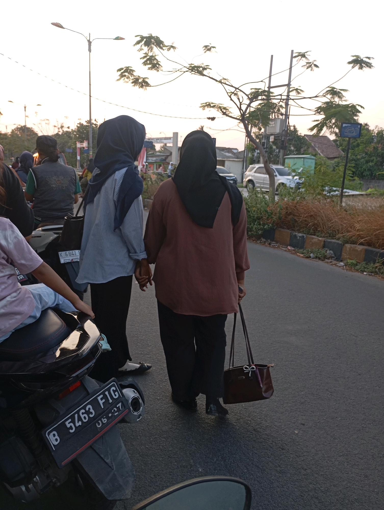 Two women are walking hand-in-hand on a street in Babelan, Indonesia. The woman on the left wears a blue and white striped long-sleeved shirt and black pants, holding a black handbag. The woman on the right wears a brown long-sleeved shirt and black pants, also holding a brown handbag adorned with a bow. A motorcycle with license plate "B 5463 FIG" is parked on the left. In the background, trees, houses, a streetlight, and other vehicles are visible. The sky is overcast.