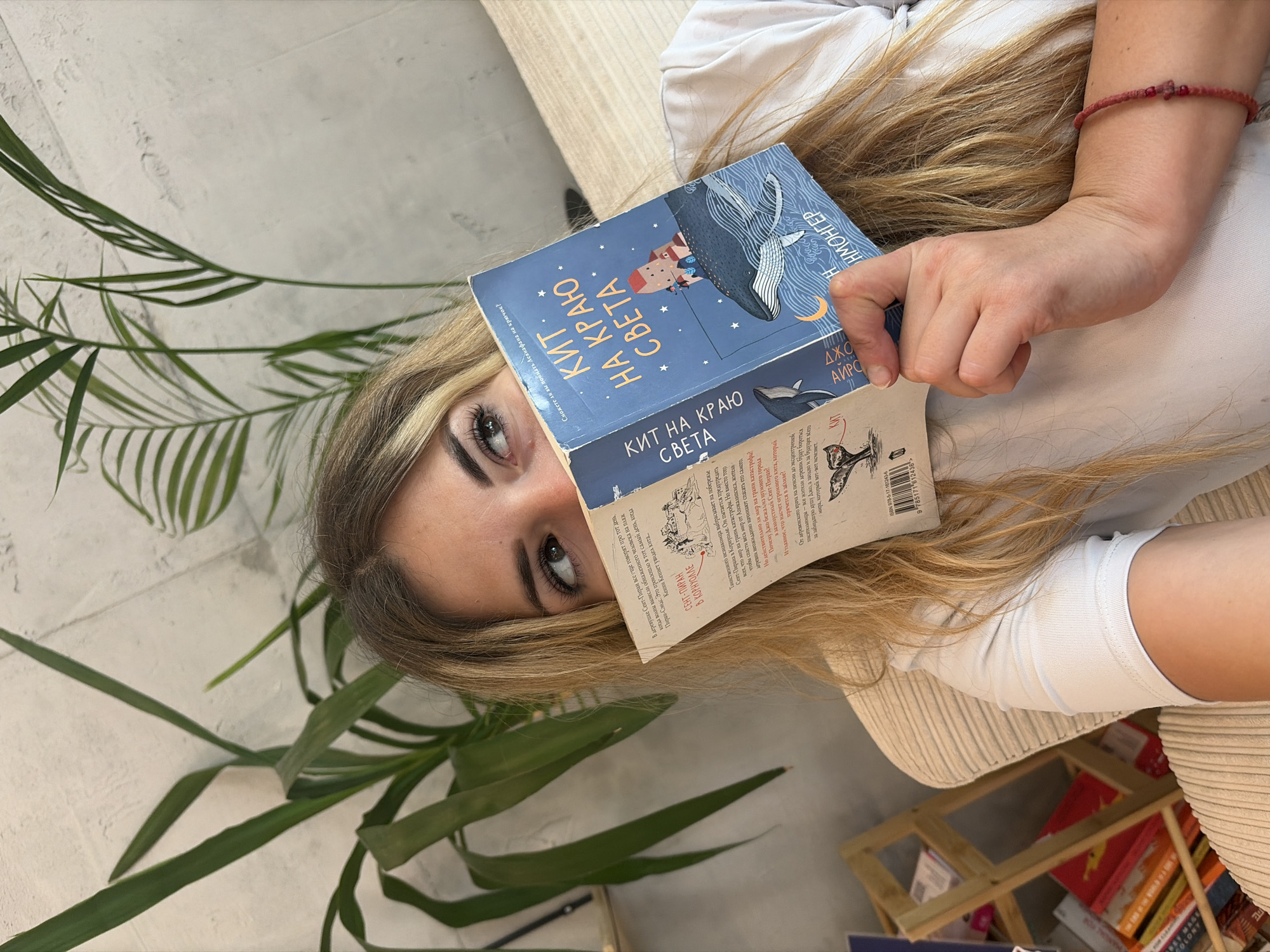 A young woman with blonde hair is lying down, holding a book in front of her face, with only her eyes visible above the cover. She is wearing a white t-shirt and a red beaded bracelet on her left wrist. The book she is holding is blue and features an illustration of a whale and a small house on its back, with stars and a crescent moon in the background. The title on the book appears to be in Russian and reads "КИТ НА КРАЮ СВЕТА" (Kit na krayu sveta), which translates to "The Whale at the End of the World." She is partially obscured by the book and large green plants with long, thin leaves that are in the foreground and to her left. Behind her, a textured, light-colored wall is visible. To the left of the woman, a wooden shelf unit filled with books can be seen. The scene appears to be indoors, possibly in a cafe or a bookstore. There is no discernible indication of the city of Budva, Montenegro.