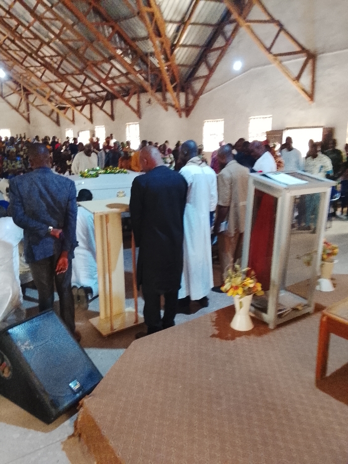 The image shows a large gathering of people in an indoor setting with a wooden, vaulted ceiling and light brown tiled floors. The people are assembled in the middle of the room. There are several rows of wooden pews that are partially visible in the background. The main focus of the gathering is a wooden podium, behind which a man in a black suit is standing, bowing his head. There is a man in a white robe standing behind him with his arms crossed, and a third man in a black suit, standing further back, also bowing his head. Directly in front of the men, there is a large, glass case that is covered with a red cloth, inside which a religious object may be displayed. The case is surrounded by several floral arrangements. At the front of the image, there is a large, black speaker on the floor. The scene is likely a religious ceremony in Mukaiya, Nigeria, located at coordinates 8.4 latitude and 8.343 longitude.