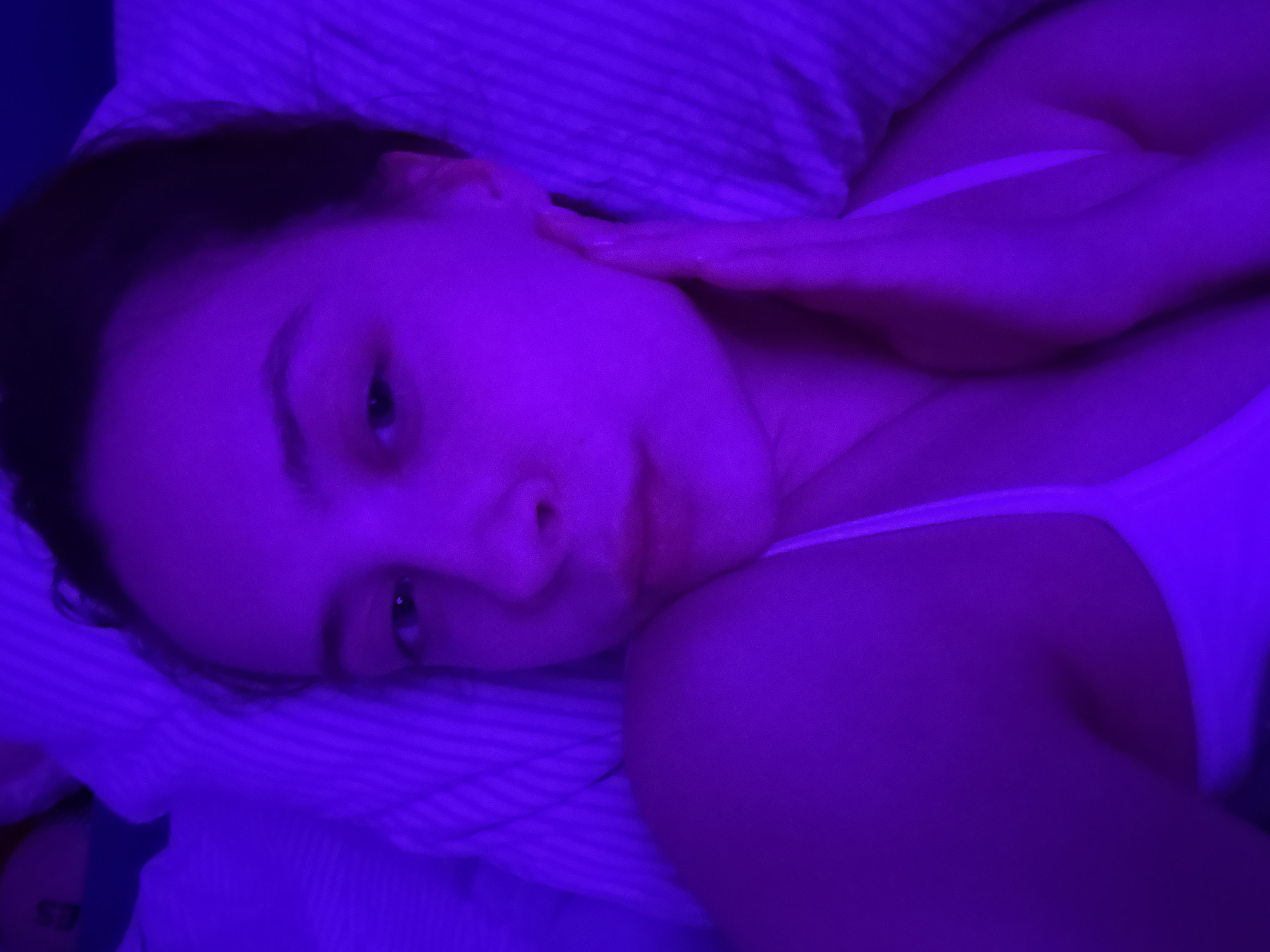 A woman is lying in bed in Прикамский, Russia. She is wearing a white tank top and has her hand on her cheek. The room is lit with a blue light. The bed has striped sheets.