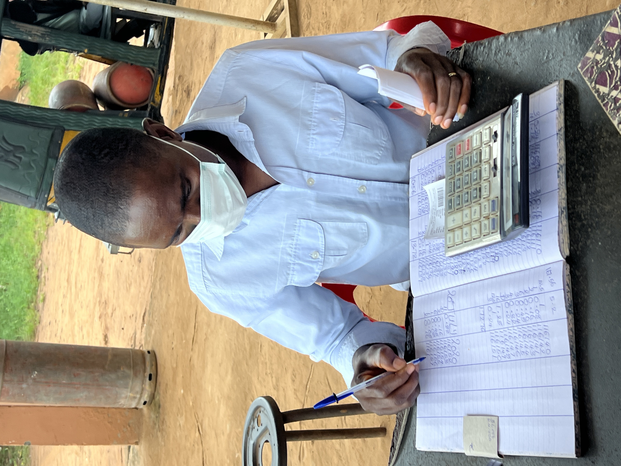 In Nkpor, Nigeria, a man wearing a light blue collared shirt and a white face mask sits at a dark table, writing in a ledger. He holds a blue pen in his right hand and a small stack of white paper in his left. The ledger is open, showing columns of handwritten numbers. A silver calculator rests on top of a separate piece of paper on the ledger. In the background, a large metal cylinder, possibly a gas tank, stands near a sandy area. To the right, part of a vehicle with visible gas tanks and a person seated inside is present. The environment appears to be outdoors with green vegetation in the distance.