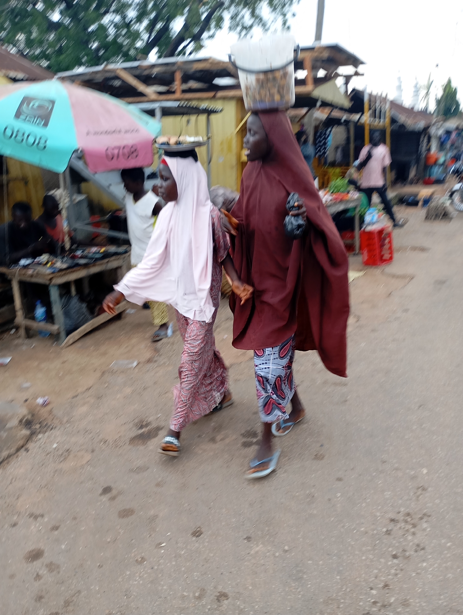 In Shendam, Nigeria, a street scene unfolds with two women walking. The woman on the right, dressed in a maroon abaya, carries a white bucket on her head and a dark bag in her right hand. The woman on the left wears a light pink hijab and a patterned dress. They are in a marketplace environment with stalls and various goods visible. An umbrella with "ERIN" and "0808" printed on it is prominent in the background. The ground is a dusty, unpaved surface. The image is blurred, suggesting motion.
