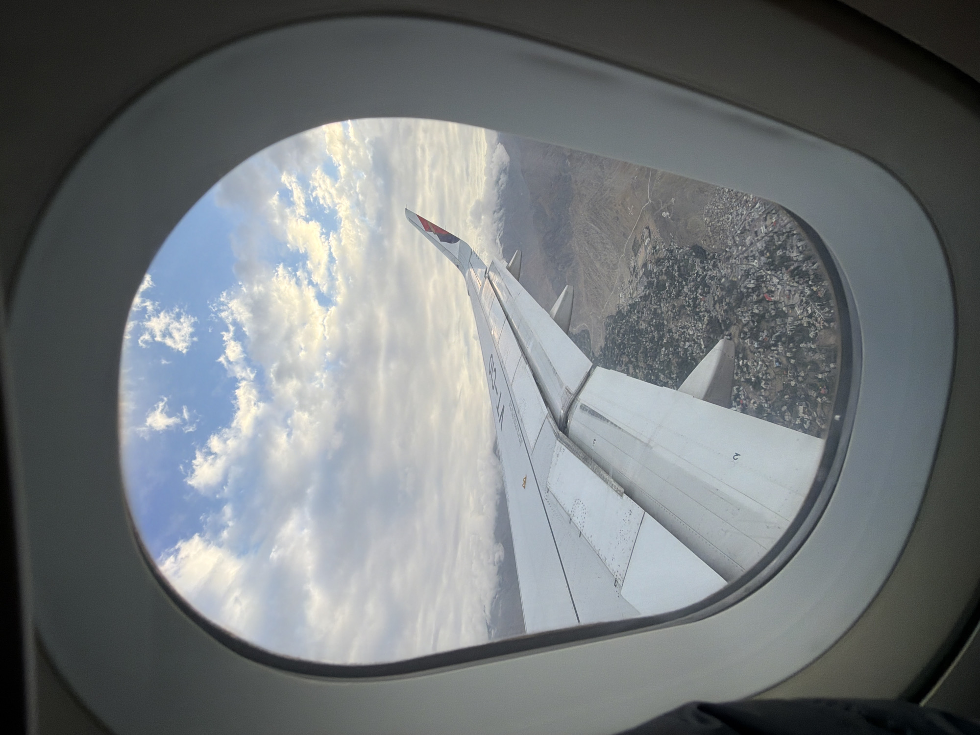 The image is taken from an airplane window, showing a view of the city of Leh, India, from above. The perspective is from an aircraft wing, extending into the frame from the lower left and middle, with the aircraft's passenger cabin window framing the scene. The wing is predominantly white and shows visible rivets and panel lines. The aircraft's tail fin, featuring red and blue accents, is visible at the tip of the wing.
Below the aircraft, the city of Leh is spread out across a mountainous terrain. The urban area appears densely populated with buildings, predominantly light-colored, clustered together. Roads are visible winding through the settlement and into the surrounding landscape. The mountainous topography is rugged, with slopes and valleys evident.
The sky above is filled with a mix of clouds. In the upper portion of the frame, a clear blue sky is interspersed with fluffy white clouds. Closer to the horizon, the clouds are thicker and more overcast, partially obscuring the distant mountain peaks. The lighting suggests it is either early morning or late afternoon, with a diffused light illuminating the scene. No people are visible in the image.