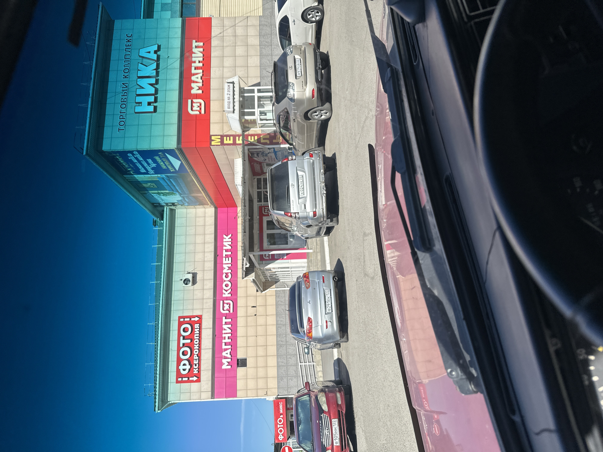 The image captures a street view in Novoaltaysk, Russia, featuring a shopping complex with prominent signage. The complex has a light-colored facade adorned with signs for "ФОТО | КСЕРОКОПИЯ" (PHOTO | XEROCOPY) and "МАГНИТ КОСМЕТИК" (MAGNET COSMETIC). Above this, a large turquoise sign reads "ТОРГОВЫЙ КОМПЛЕКС НИКА" (SHOPPING COMPLEX NIKA). Further right, a red sign with a white "M" logo states "МАГНИТ" (MAGNET). Several cars are parked in front of the complex, including a red sedan with license plate "B 105HH 122", a silver Mitsubishi Lancer with license plate "B291EK 122", a silver Toyota Land Cruiser Prado with license plate "K 024VK 22", and a gold Lexus RX with license plate "M979CB 22". A white sedan is partially visible on the far right. The sky is clear and blue, suggesting a sunny day. The photo appears to be taken from inside a vehicle, looking out through the windshield.