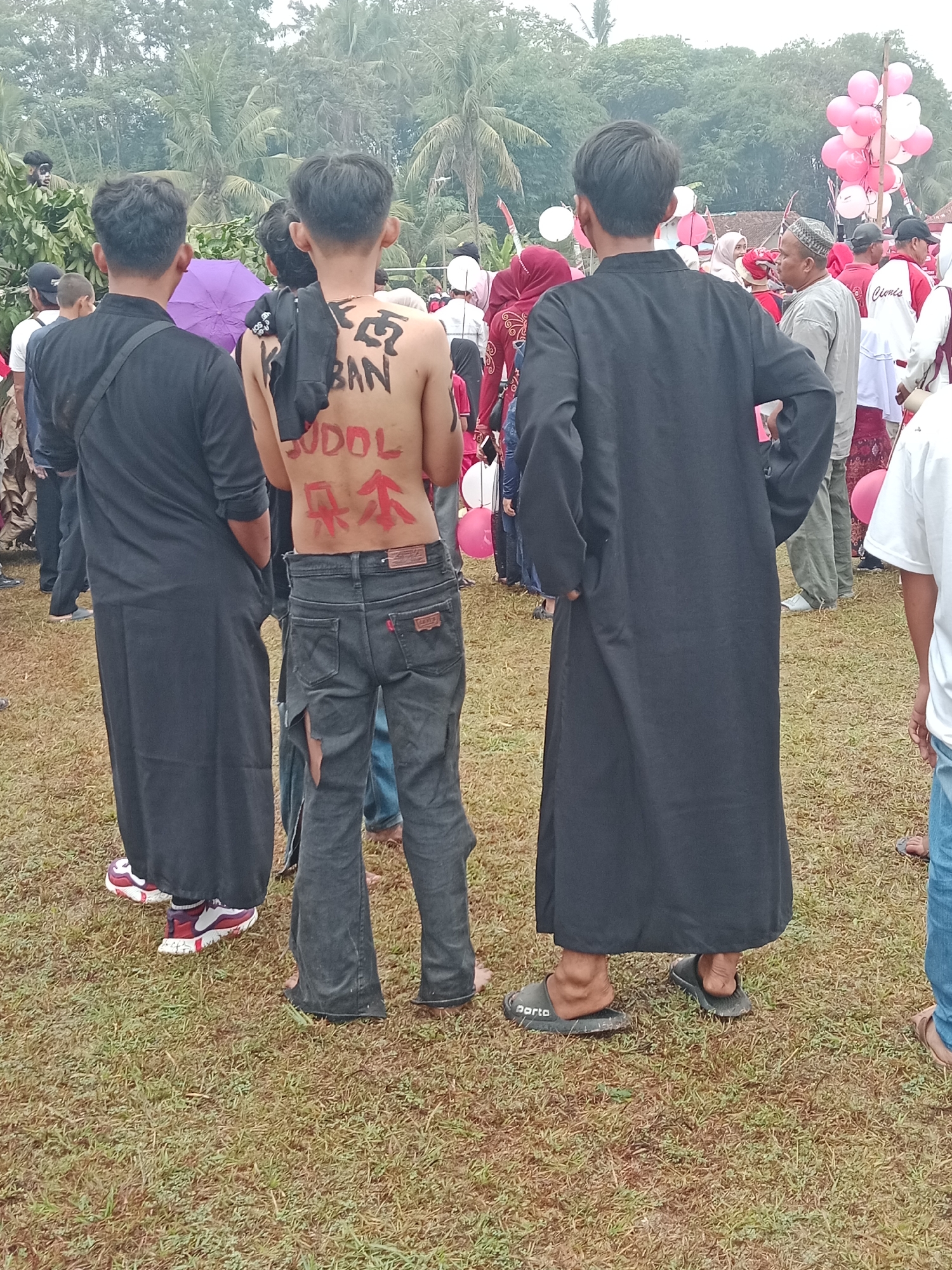 The image depicts an outdoor gathering in Bojongmengger, Indonesia, with a focus on three individuals in the foreground. The person in the center, facing away from the camera, has writing in black and red paint on their bare back. The writing appears to be in a mix of Latin and possibly Asian characters. They are wearing ripped dark denim jeans. To the left of this individual is another person wearing a long black robe and a black bag slung across their shoulder. They are also wearing athletic shoes. To the right of the central figure is a third person, also wearing a long black robe, with their hands in their pockets. They are wearing dark sandals.

In the background, a crowd of people are visible, some wearing traditional Islamic attire such as hijabs and skullcaps. There are also balloons, some pink and white, tied together and floating in the air. The environment appears to be a grassy outdoor area, with trees and foliage visible in the distance, suggesting a rural or semi-rural setting. The overall atmosphere suggests a community event or celebration.