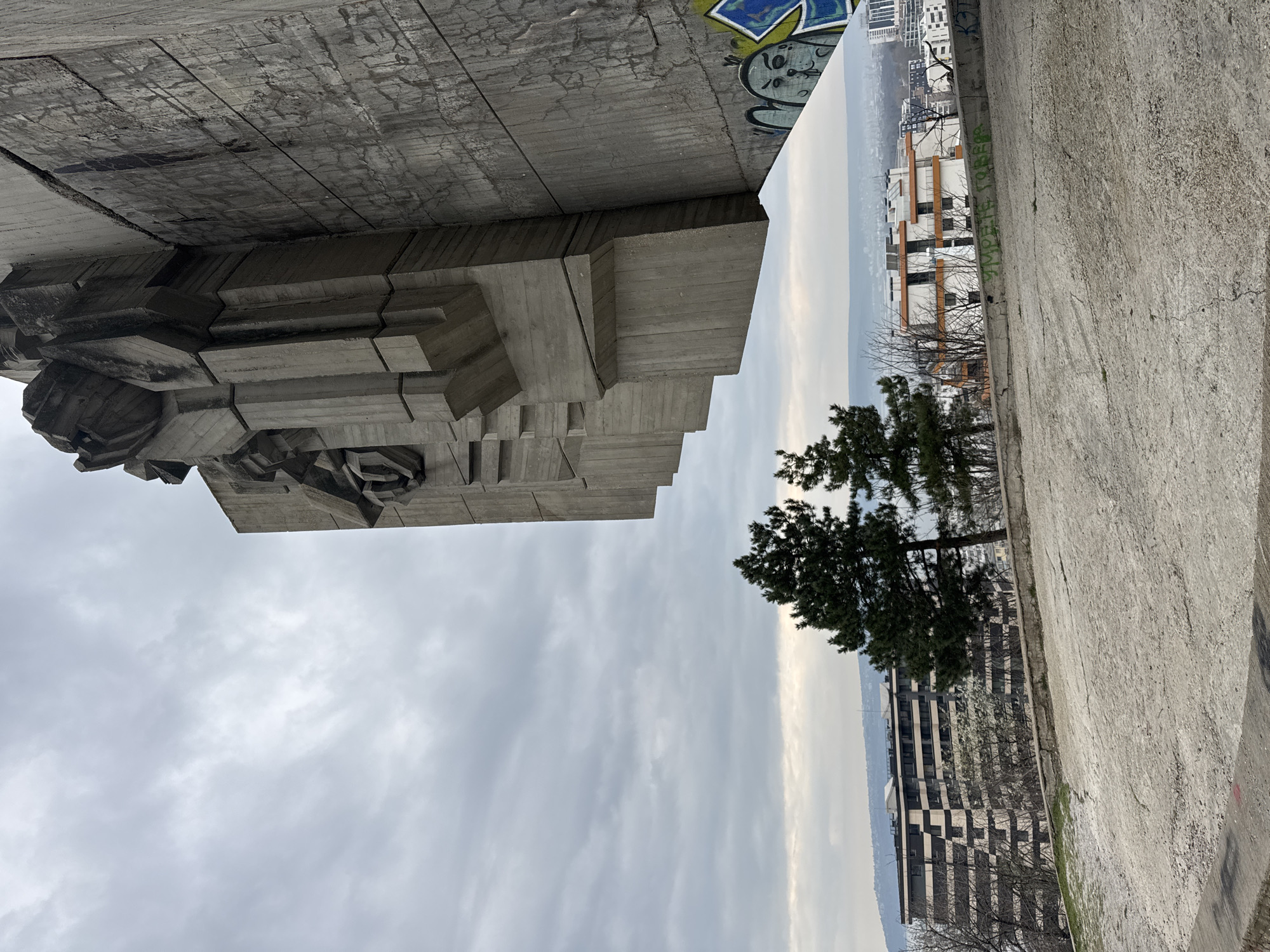 The image shows the Monument of the Soviet Army in Varna, Bulgaria, from an unusual perspective, rotated counterclockwise. The monument is a massive concrete structure with abstract relief sculptures. Graffiti is visible on the upper part of the monument. In the background, buildings are visible in the city, partially obscured by a tree. The sky is overcast. The foreground features a concrete walkway or ledge.