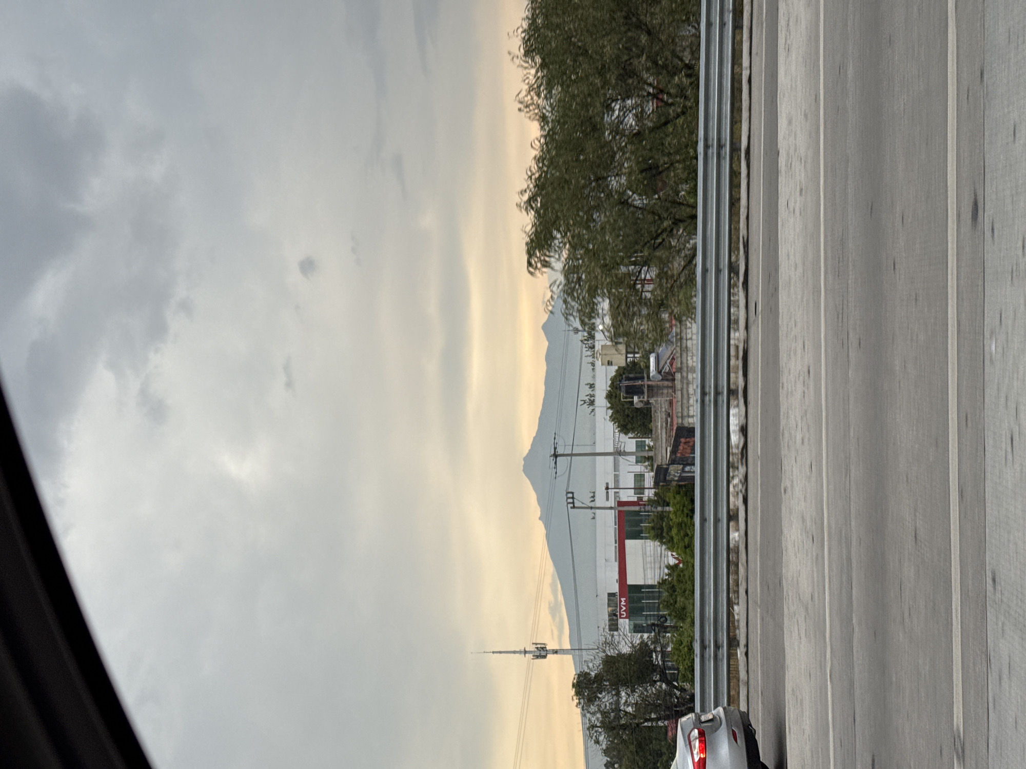 The image, taken from a moving vehicle, shows a view of San Bernardino Tlaxcalancingo, Mexico. In the foreground is a multi-lane road with a metal guardrail. A partial view of a light-colored car is visible in the lower right corner. Beyond the guardrail and road, a low mountain range is visible against a cloudy sky. In front of the mountains are buildings, trees, and what appears to be a radio or communications tower. One visible building is labeled "UVM" with red lettering. The sky is overcast with varied shades of gray and a faint yellow tint near the horizon.