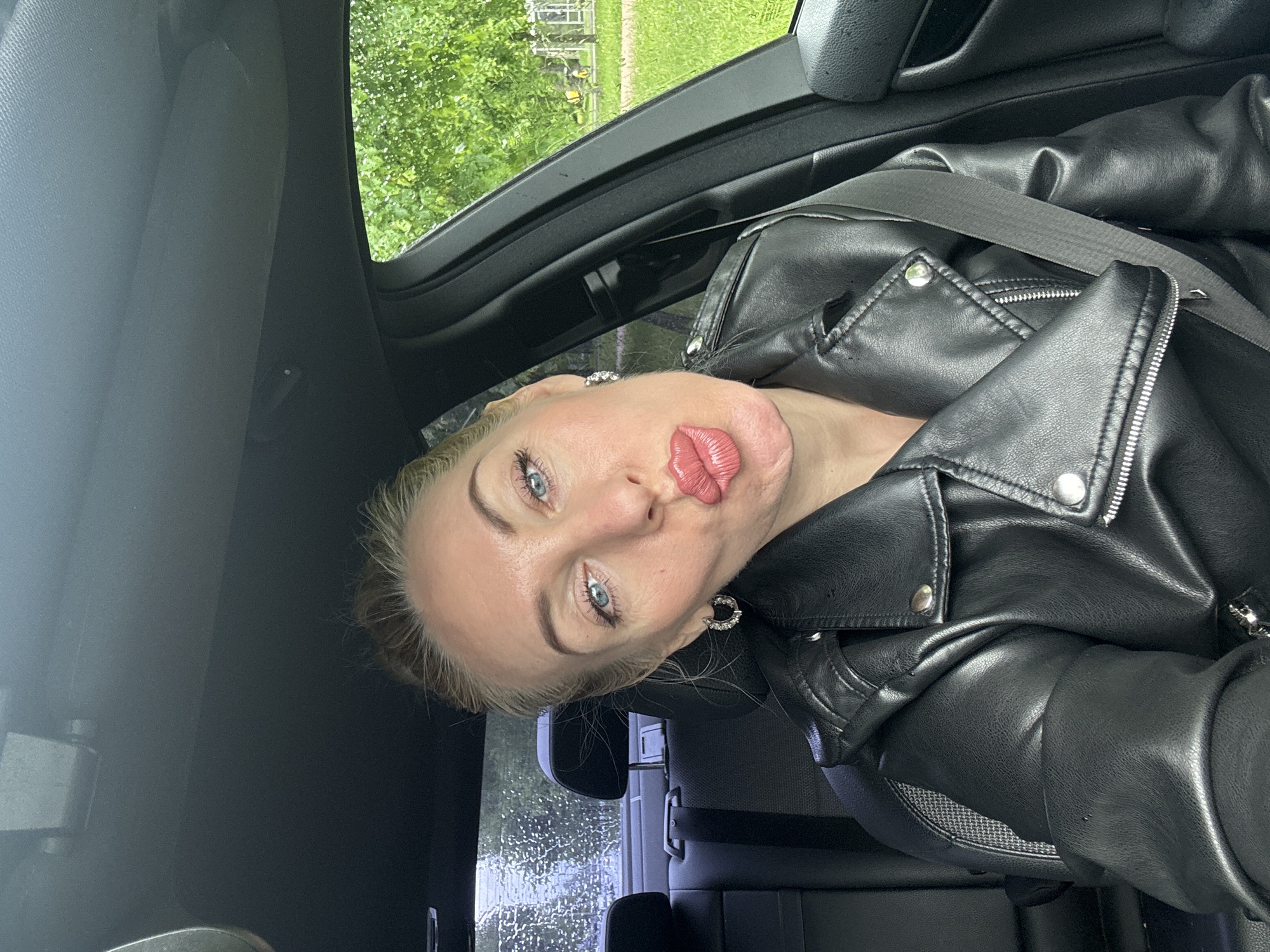 A person is inside a vehicle, likely a car, wearing a black leather jacket and a seatbelt. The individual is positioned in the driver's seat, and there are visible trees through the car window. The location appears to be a city area, given the buildings visible in the background, but the exact location is unknown.