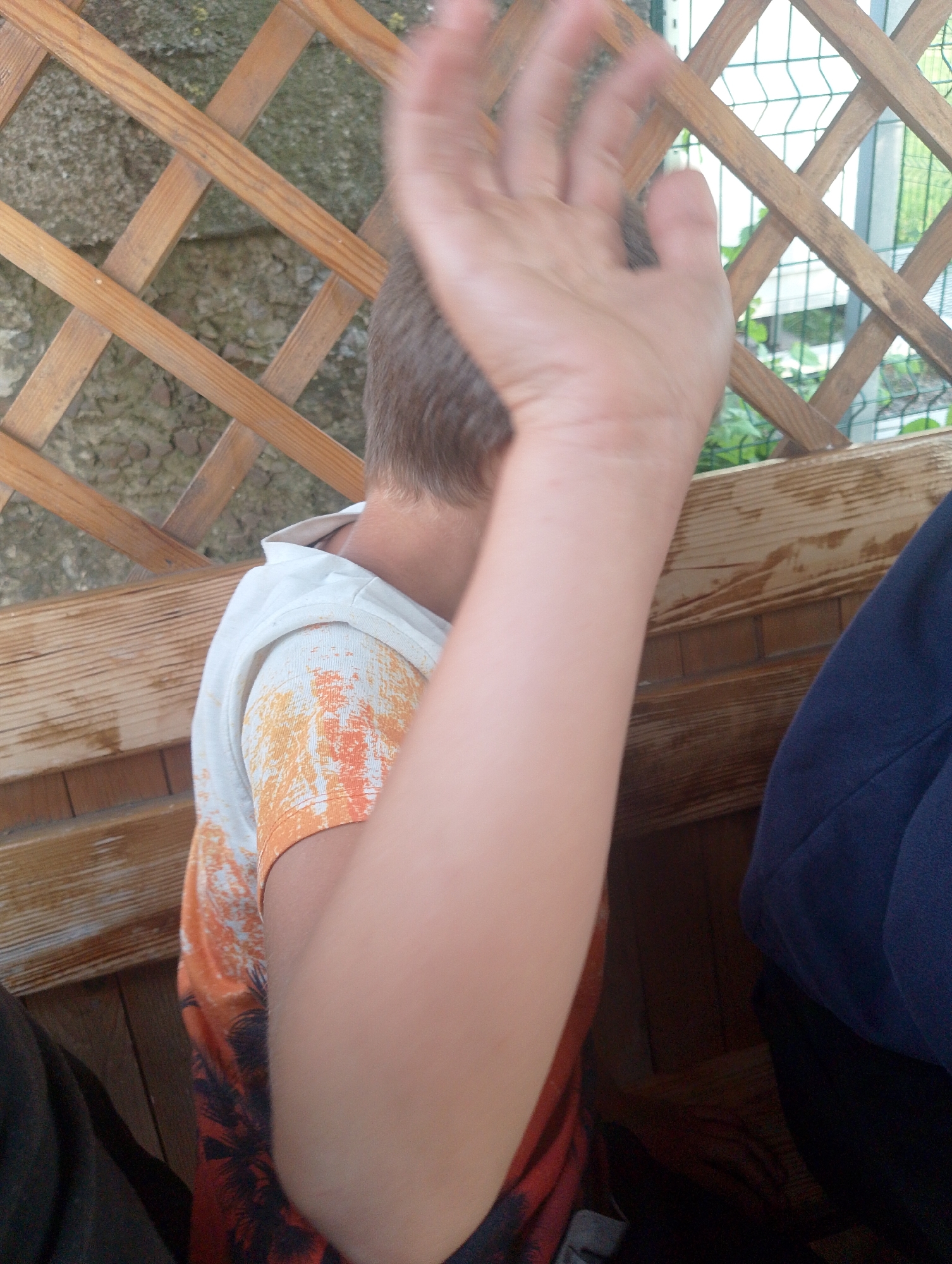 The image shows a close-up of a young person's arm and shoulder, obscuring much of their face and the surrounding environment. The person is wearing a white t-shirt with an orange and black pattern, possibly depicting palm trees or a similar motif. Their right arm is raised and blurred, suggesting movement. A dark blue jacket or shirt is visible on the right side of the frame. The background includes a wooden lattice structure and a concrete wall, hints of an outdoor setting, possibly a park or garden area. The lighting appears to be natural, suggesting it's daytime, and the overall impression is casual and candid. The specific location is Ustyvytsia, Ukraine. No text is visible in the image.