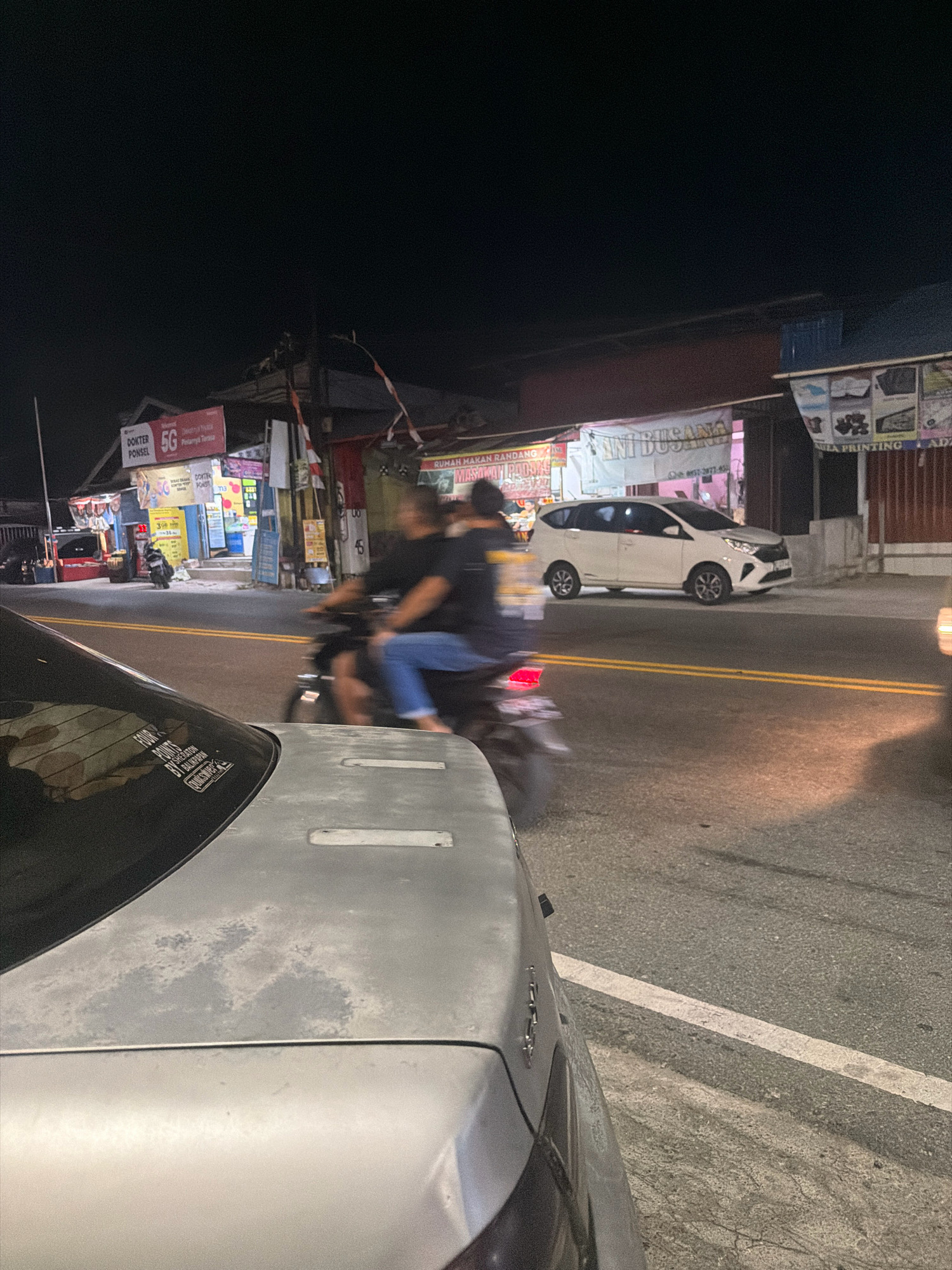 The image depicts a nighttime street scene in Balikpapan, Indonesia, likely in a commercial area with multiple shops lining the road. In the foreground, the hood of a silver car is visible, showing signs of wear and tear. The car has a sticker on its rear window that reads "FOUR POINTS BY SHERATON BALIKPAPAN." A motorcycle with two occupants is in motion on the right side of the image, with the rider blurred due to the movement. The motorcycle's taillight is illuminated red. A white car is parked on the left side of the street. Various signs are visible above the shops, including "DOKTER PONSEL 5G," "RUMAH MAKAN RANDANG," and "ANI BUSANA." The street is paved with asphalt and marked with yellow lines. Streetlights and the illumination from the shops create a dimly lit environment.