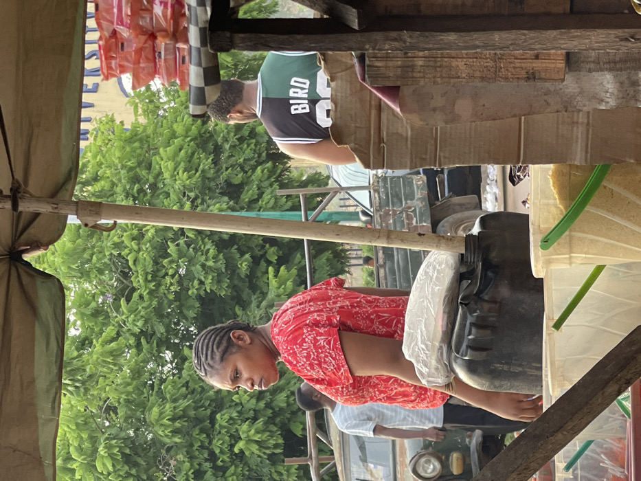 The image depicts an outdoor market or roadside stall during the daytime, characterized by a rustic setup and several individuals. The primary orientation of the image is rotated 90 degrees counter-clockwise; for this description, it will be analyzed as if correctly oriented.
In the foreground, on the left, an olive-green canvas awning provides shade, supported by visible poles. To the right of the awning, a young woman stands behind what appears to be a stall counter, made of rough wood and possibly cardboard. She is wearing a red short-sleeved top with a white patterned design and has her dark hair styled in intricate braids. Her gaze is directed downwards, suggesting she is focused on an item or task in front of her.
Immediately to her right, on the stall counter, sits a large, black plastic container, possibly a jerry can or a drum, with a white plastic bag covering part of its opening. Further to the right, a light-colored plastic bucket with a green handle and spout is visible, containing a granular, pale yellow substance, likely grains such as rice or maize.
In the midground, behind the stall, another person, a man, is partially visible. He is wearing a green, black, and white basketball jersey with "BIRD" and the number "33" clearly visible on the back. His back is mostly turned towards the viewer, and he has short, dark hair or a beard. He is standing behind a cardboard barrier, possibly another part of a stall or a display.
The background is dominated by lush, green foliage from numerous trees, indicating an outdoor, possibly natural, setting. Behind the woman and the black container, a portion of an older, dark green utility vehicle, possibly a truck or jeep, can be seen, with its circular headlight visible. Further into the background, other indistinct figures and white objects, possibly containers or goods, are faintly visible.
Above the trees and to the far left (upper left when rotated), several small, orange-colored packaged items are hanging, likely products for sale. A black and white checkered pattern is visible on a vertical pole or structure near these hanging packages. Above these, a light-colored sign with some faint blue text is also present.
The overall impression is that of a bustling local market or a community gathering point, with people engaging in commerce or daily activities under natural daylight.