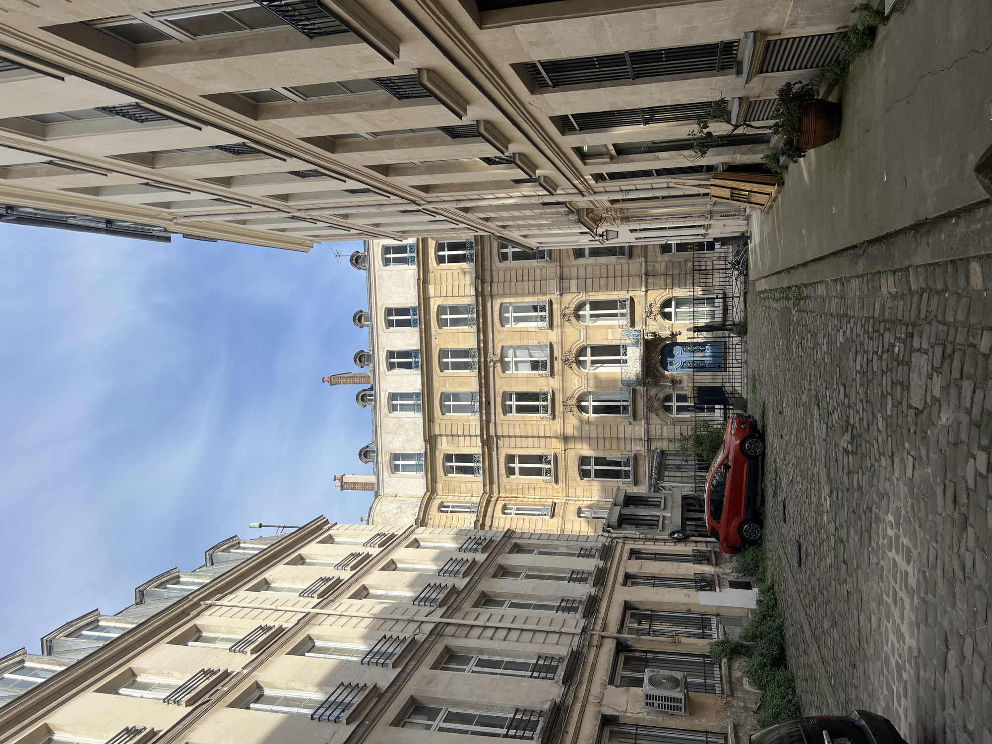 A narrow Parisian street scene shows buildings with light beige stone facades, multiple stories high, featuring windows with dark metal railings and traditional architectural details like curved cornices. A red compact car is parked along the right side of the cobblestone street. A section of blue sky is visible between the tops of the buildings. The architecture and street style are consistent with the city of Paris, France.