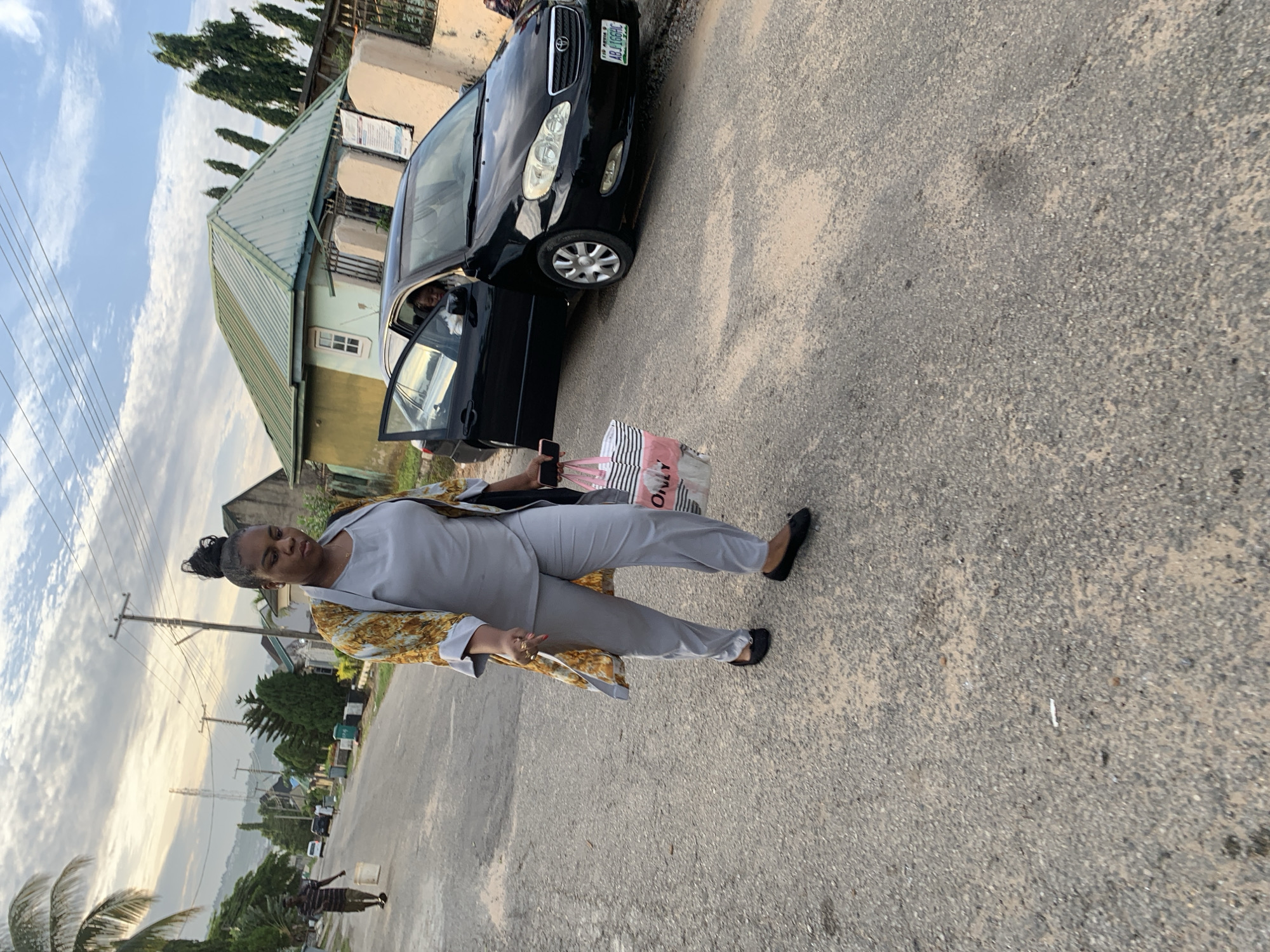 A woman is walking on a paved road in front of a black sedan with its driver's side door open. She is wearing a gray outfit and a patterned open-front jacket. She is carrying a striped bag with pink and white stripes and a pink object inside, and a phone in her right hand. A man carrying a white bucket is walking away from the camera on the left side of the road. Houses and vegetation are visible in the background. The scene appears to be in Abuja, Nigeria.