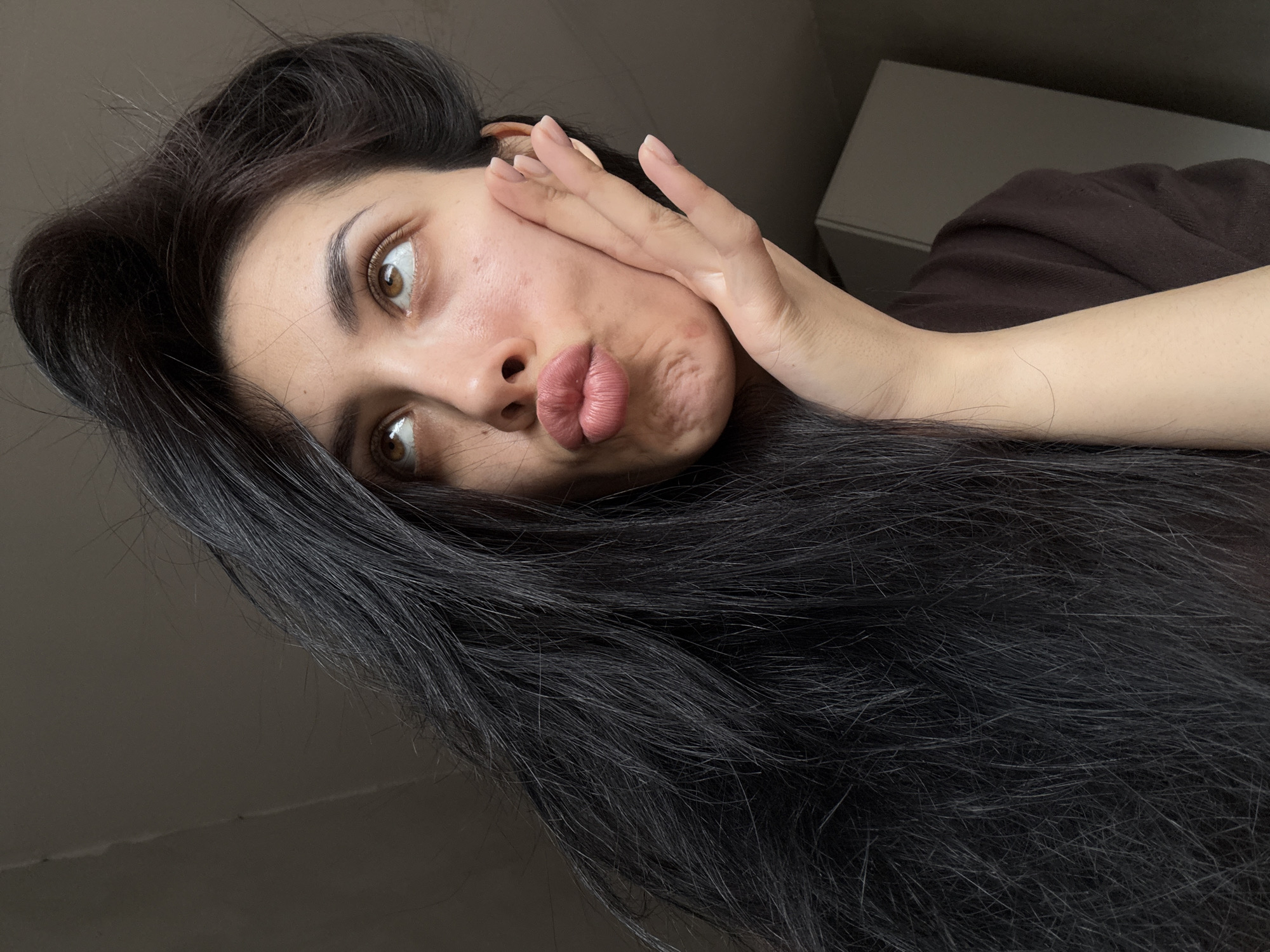 A young woman is captured in a close-up, angled selfie, likely taken indoors in Votkinsk, Russia. Her head is tilted, with her long, dark, wavy hair flowing down the left side of the frame and across her chest. She has light brown eyes, which are wide and gazing towards the upper right, and she is making a distinctive pouty face with her full, pink lips. Her left hand is raised to her left cheek, with her manicured fingers visible. Her complexion is fair, showing some natural skin texture and minor blemishes, including a small reddish mark on her cheek. She is wearing a dark brown top, which is partially visible. The background is simple and minimalist, featuring a light-colored wall and a dark, indistinct object or piece of furniture behind her right shoulder. The lighting is soft and even, typical of an indoor setting. No text is visible in the image.