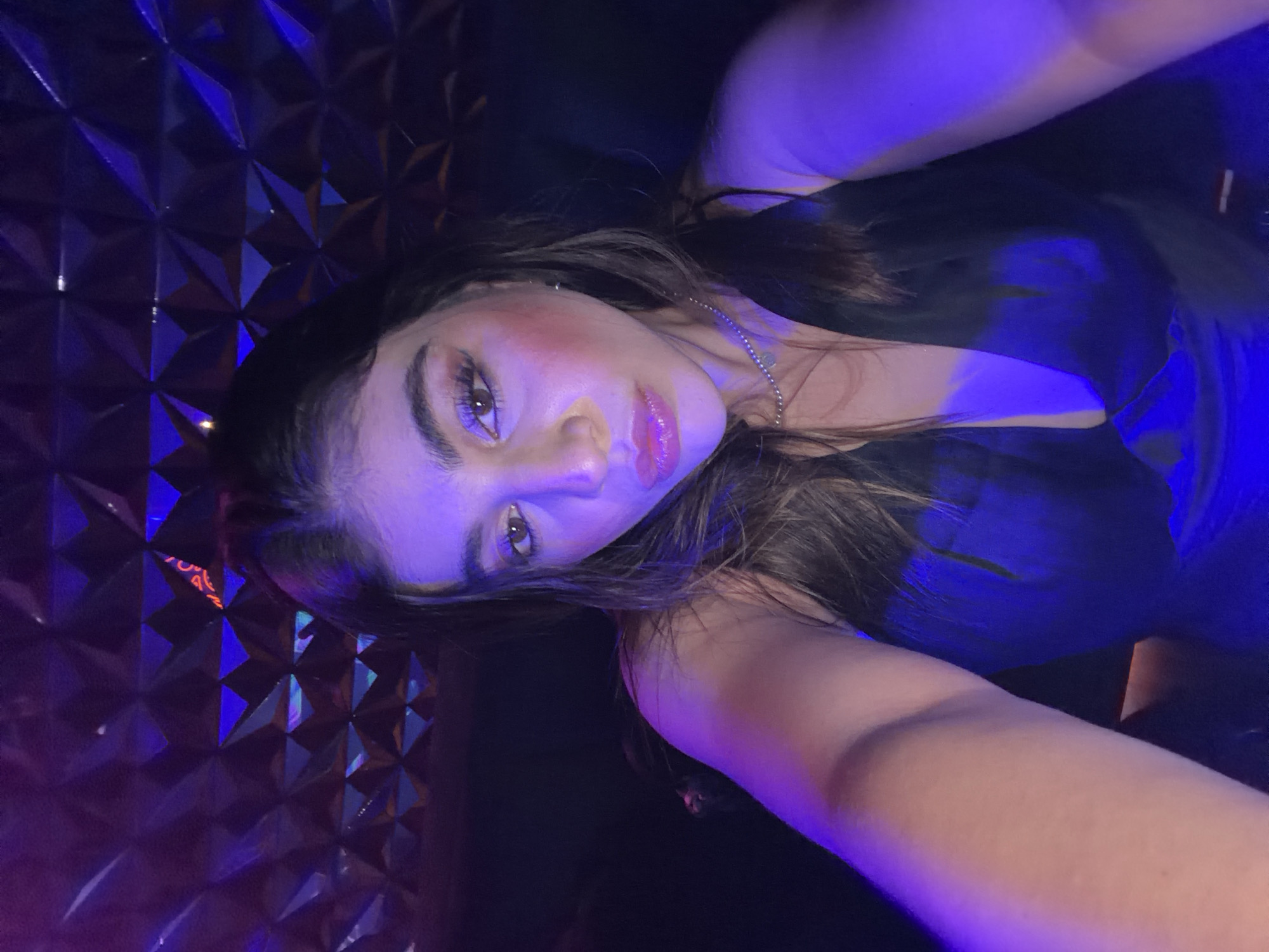 A young woman with long dark hair takes a selfie, illuminated by blue and purple lighting. She wears a dark-colored top, and a delicate silver necklace is visible. The background features a textured, geometric design, possibly a wall panel. The image's location is not discernible from the available visual information, and no information about the location in San Bernardino Tlaxcalancingo, Mexico can be provided based on the image.